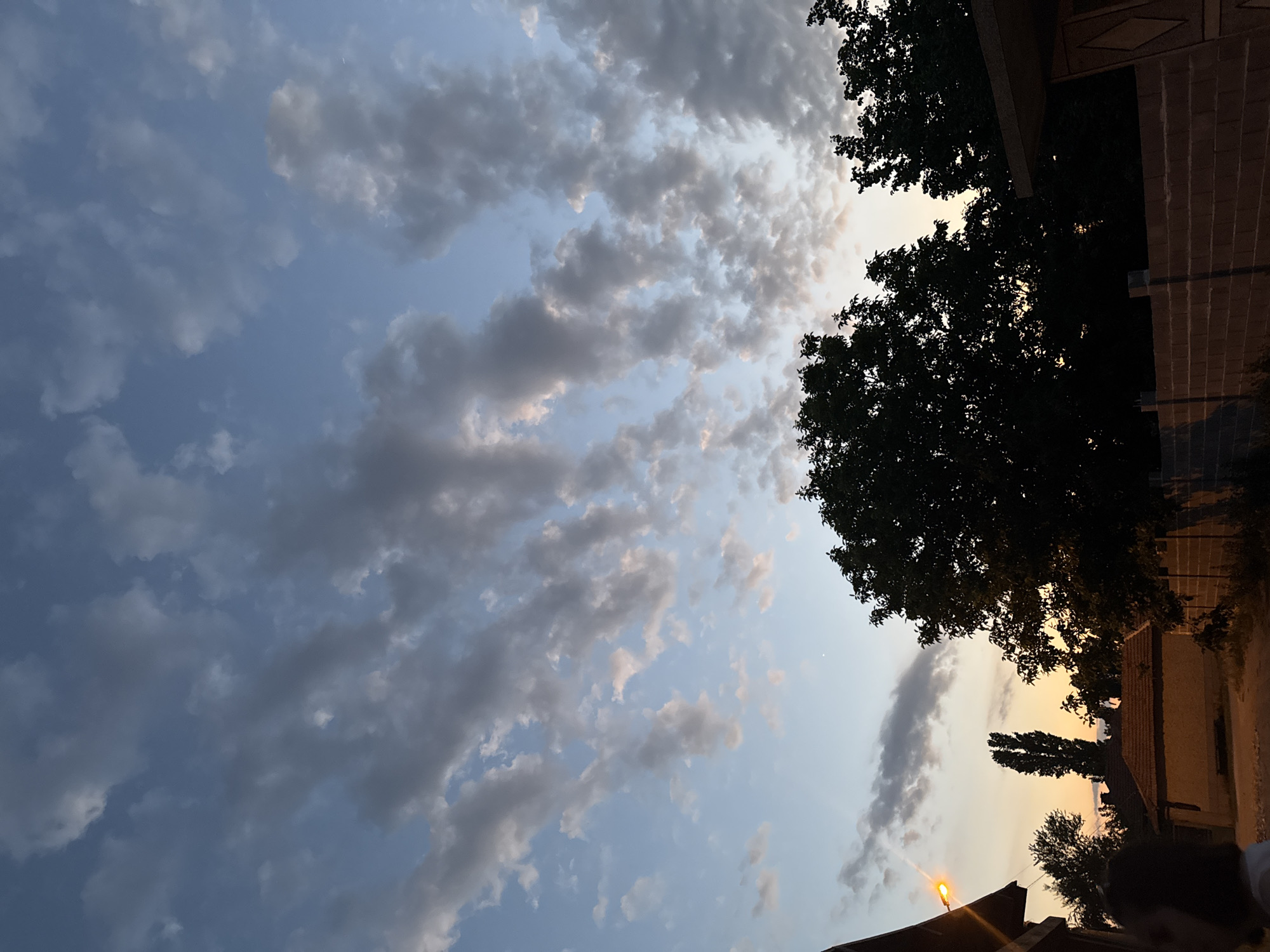 The image shows a view of the sky with clouds and silhouettes of trees and buildings. The sky is a pale blue with scattered gray and white clouds, some tinged with pink and orange from the setting sun. To the right, dark green tree canopies are visible, along with the corner of a building with a brick facade and a tiled roof. A street lamp with an orange light is also visible in the lower left corner, suggesting it is dusk. The overall mood is peaceful and serene, characteristic of a late evening in a rural or suburban setting. The location is Topolchane, Bulgaria. There is no visible text in the image.