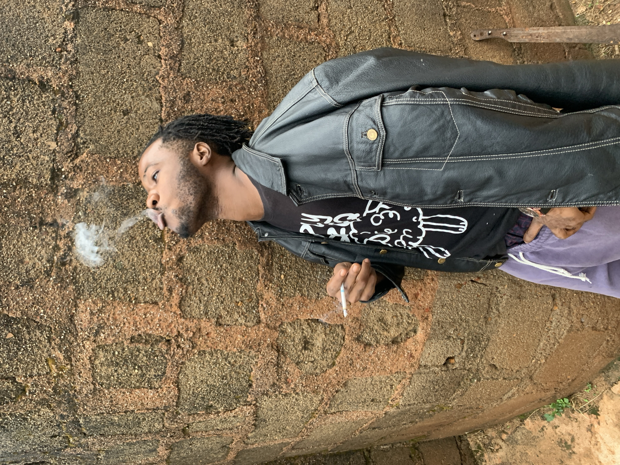 A young Black male with dreadlocks is leaning against a rough, textured brick wall, exhaling smoke from a cigarette held in his right hand. He is wearing a black t-shirt with a white graphic design, a black leather jacket with white stitching, and light purple sweatpants. The wall is made of large, irregularly shaped bricks, some reddish-brown and others a lighter, sandy color, with visible mortar. The setting appears to be outdoors, with a hint of green vegetation visible at the bottom left, suggesting an urban or semi-urban environment in Jos, Nigeria. A rusty metal object, possibly a machete, leans against the wall to the right of the subject. The lighting is natural, casting subtle shadows.