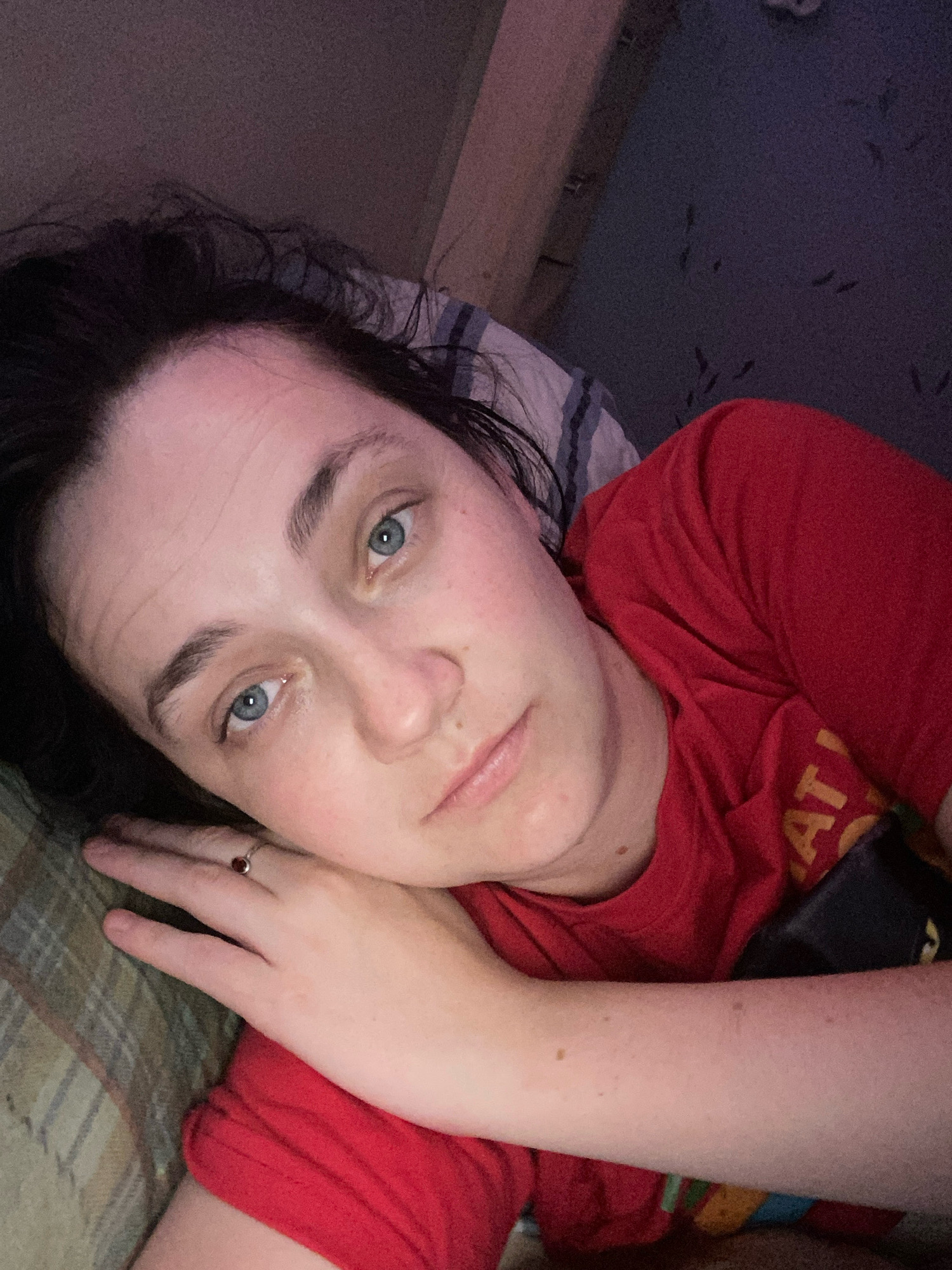 A close-up, overhead, selfie-style shot shows a person with dark hair and blue eyes lying on their side in bed. The person is wearing a red t-shirt. The left arm is bent at the elbow and resting near the person's face. A ring with a dark stone is on the ring finger of the left hand. The background appears to be a wall with a subtle pattern. The lighting is dim, suggesting it is nighttime or early morning. The image does not provide any information about a specific city or location.