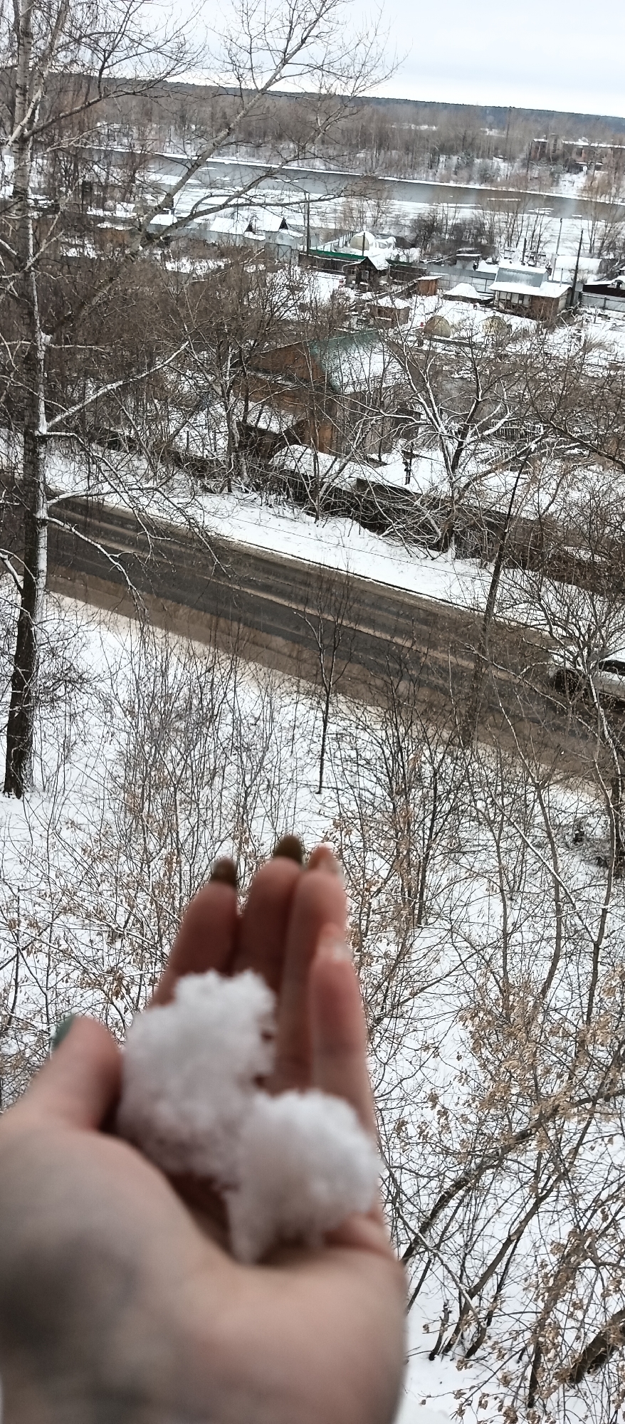 A high-angle shot shows a hand holding a clump of snow, with a winter landscape in the background. The background includes bare trees, a road, scattered houses, and a distant river or body of water. The location appears to be a city or town during winter.