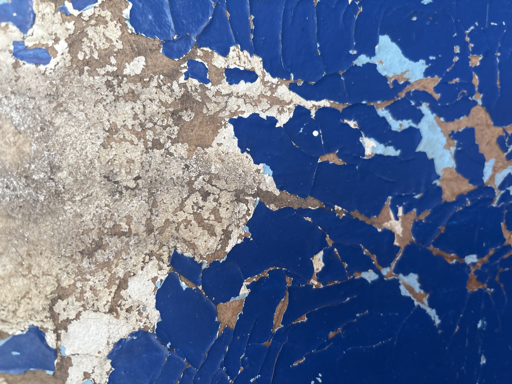 A close-up, abstract shot of peeling blue paint revealing a textured, light-colored surface underneath, possibly concrete or wood. The paint is cracked and flaking in irregular patterns, with areas of lighter blue and brown showing through.