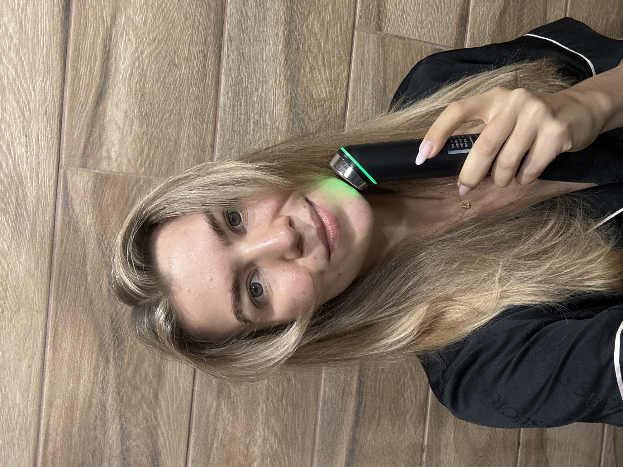 A young woman with long, blonde hair is pictured indoors, holding a black skincare device to her right cheek. The device has a metallic silver tip and emits a bright green light onto her skin. She is looking directly at the camera with a neutral to slightly soft expression. Her eyes are light-colored, and she has a few minor blemishes visible on her cheek.
She is wearing dark, possibly black, clothing that appears to be made of a silky or satin-like fabric, possibly pajamas, with thin white piping along the edges of the collar or lapel. The fabric of her clothing has a faint, repetitive pattern of dark grey text, which is difficult to read but seems to include letters like "S" and "ECR". Her fingernails are manicured with a light pink or nude polish. A delicate gold necklace with a small, heart-shaped pendant is visible around her neck.
The background consists of light brown wooden panels, arranged vertically, suggesting either a wall or a wooden floor used as a backdrop. On the skincare device, near where her fingers are, there is a small screen or label with unreadable text that includes what appears to be Korean characters and some faint English numbers. The image is rotated counter-clockwise.