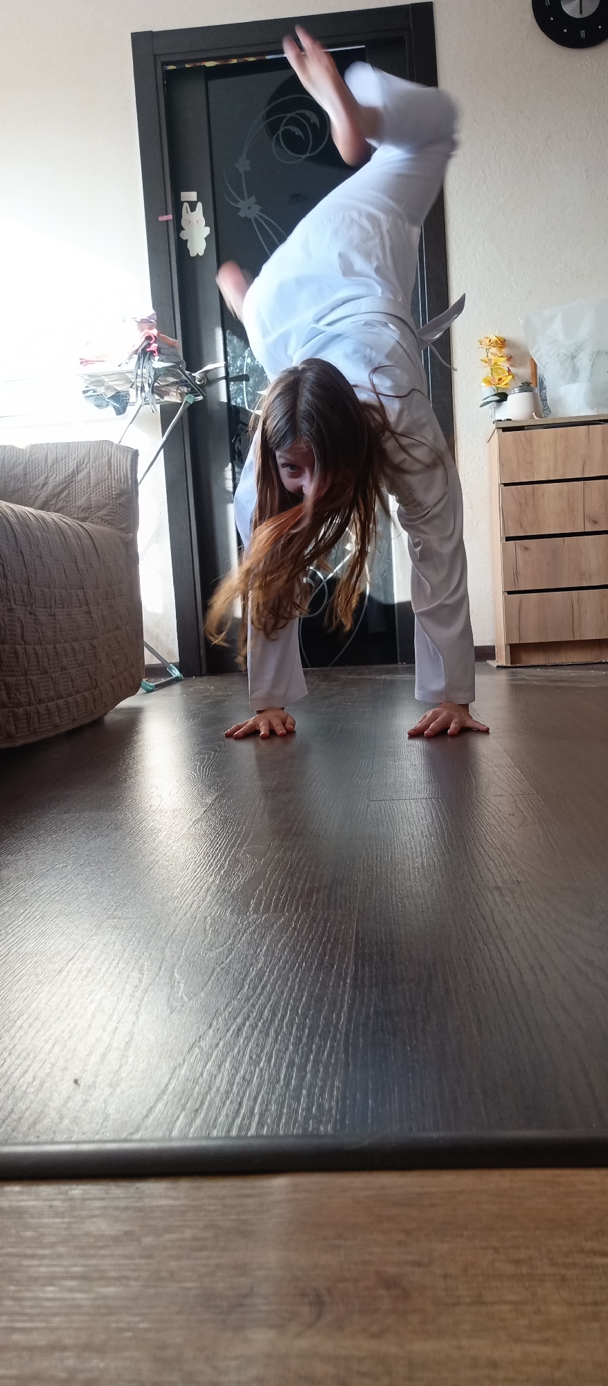 A young person in a white martial arts uniform with a black belt is performing a handstand indoors. The person's feet are elevated and slightly out of focus. Their hands are planted on a shiny, dark wooden floor. Their brown hair is hanging down toward the floor. Behind the person is a dark wooden door with a white rabbit sticker on it, a tan couch, an ironing board, a wooden dresser with yellow flowers on top, and a clock on the wall. The location appears to be a domestic setting. The city and specific location are unknown.