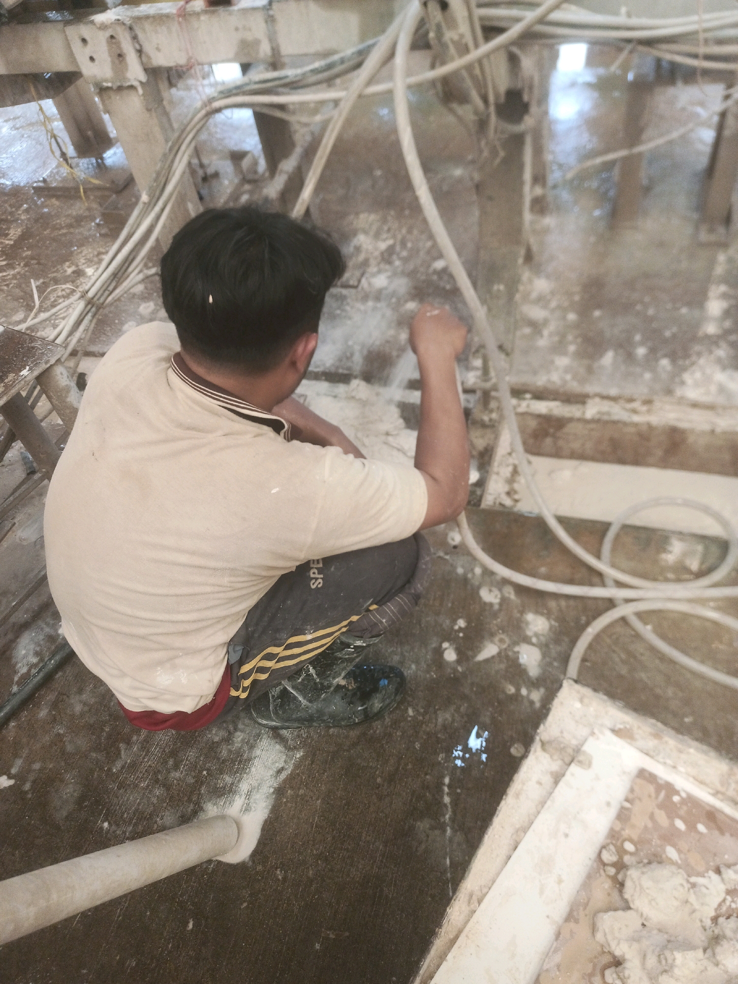 A person is working in an industrial setting in Lebaniwaras, Indonesia. The person is wearing a light-colored t-shirt with a brown collar and dark pants with yellow stripes. They are crouched down and appear to be operating some sort of machinery that involves water and possibly dust, as indicated by the wet and dusty environment. Several white hoses are visible, along with metal structures and debris on the floor. The overall scene suggests a construction or manufacturing site.