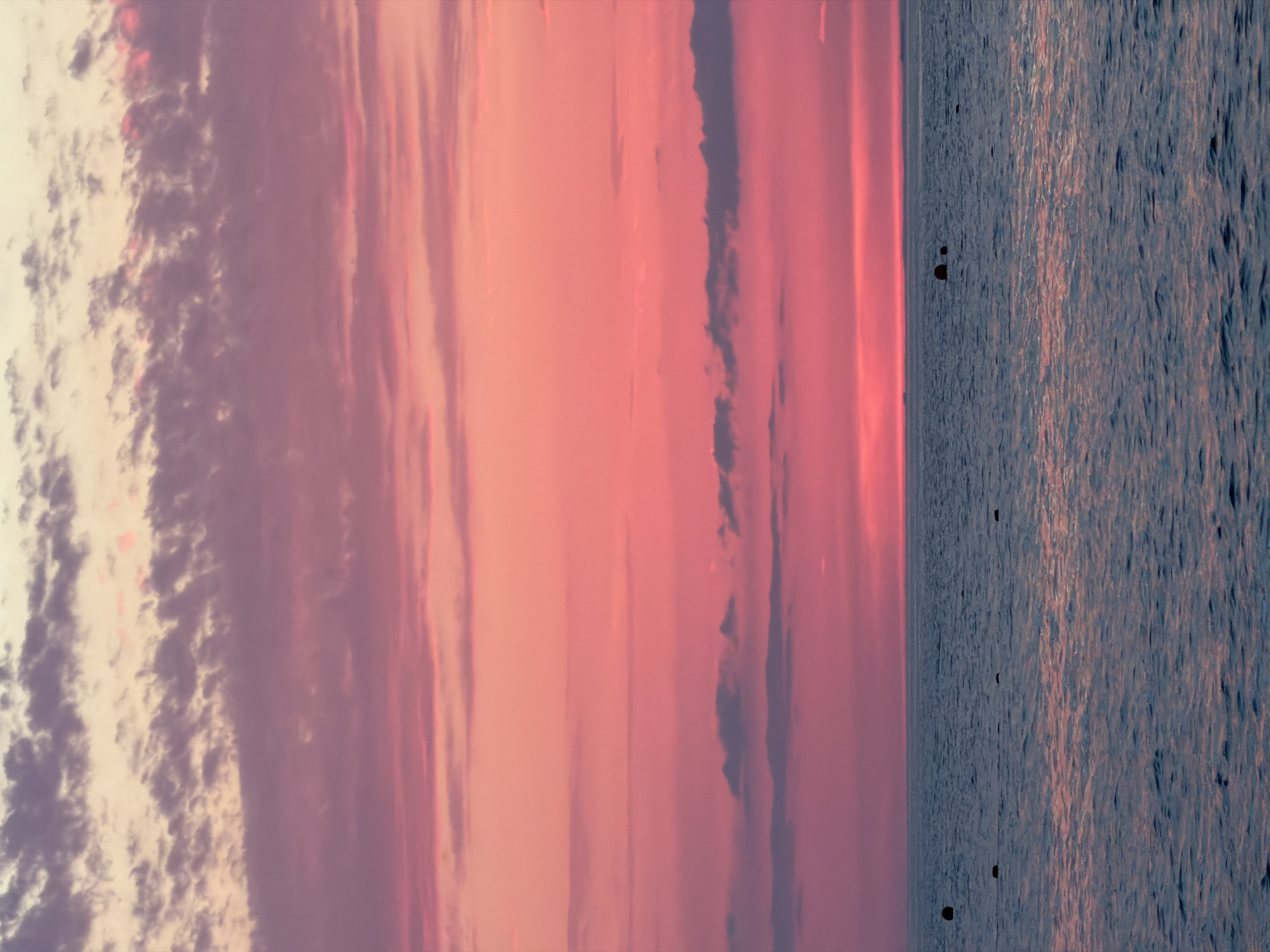 The image shows a body of water with a pink and purple sunset sky above. Several dark buoys are visible in the water. The sky is filled with layered clouds, with the lower clouds appearing pink and the upper clouds a lighter purple and cream color. The water is dark blue with gentle ripples reflecting the colors of the sky. The horizon line is clearly defined between the sky and the water.