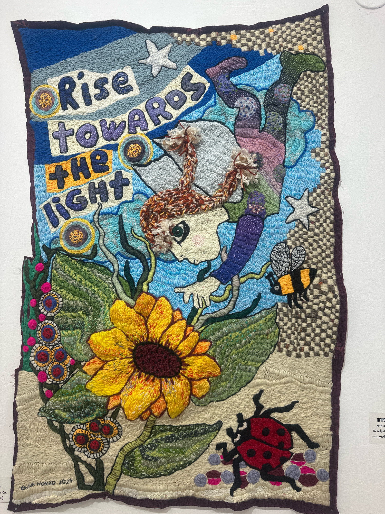 This image shows a tapestry, likely on display in a gallery or public space, in the city of Mate Asher Regional Council, Israel. The tapestry depicts a stylized, whimsical scene with a young person with auburn hair falling or floating downwards against a vibrant blue sky. Above the figure, in large, textured letters, are the words "Rise towards the light." The tapestry incorporates various textile techniques, including weaving, tufting, and embroidery, creating a rich and tactile surface.
Dominating the lower portion of the tapestry is a large, bright yellow sunflower, rendered with intricate detail. Below the sunflower, a red ladybug with black spots is prominently displayed. Smaller elements scattered throughout the artwork include stars, a bumblebee, and various circular floral or geometric motifs. The background features a mix of textured weaving patterns, suggesting a sky and earth or landscape. The tapestry is framed by a dark, textured border.
The overall impression is one of a whimsical, uplifting, and nature-inspired artwork, possibly intended to convey a message of hope, growth, and resilience.