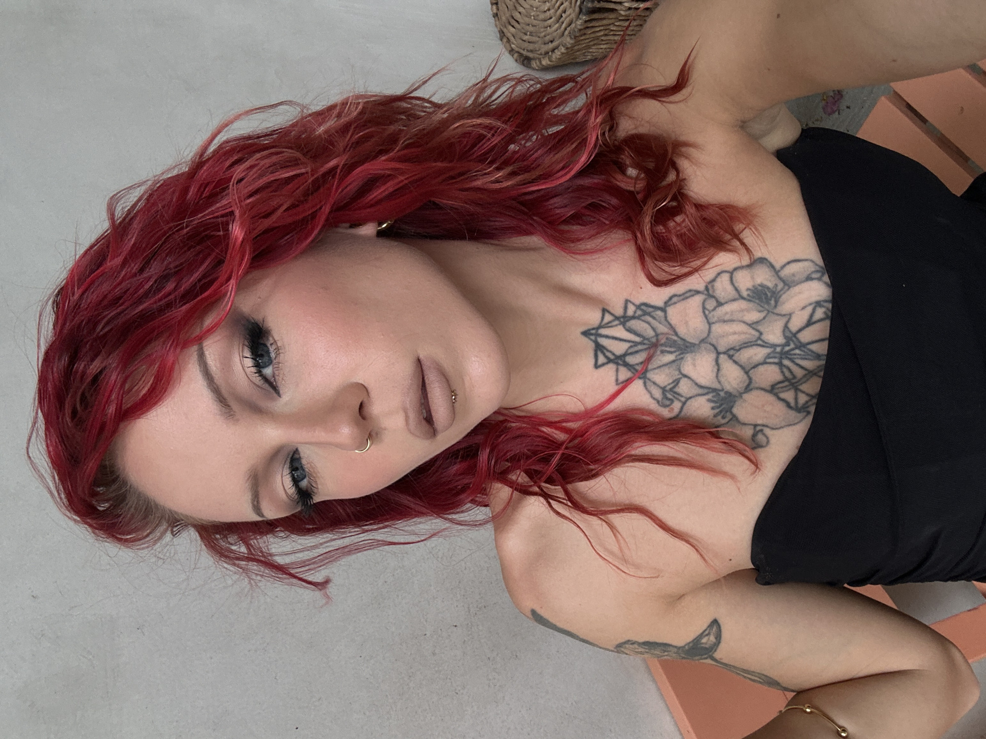 A woman with vibrant red hair is photographed from the chest up. She is wearing a black strapless top and has several tattoos visible on her chest and shoulder. Her makeup includes eyeliner, eyeshadow, and neutral lipstick. She has a nose ring and a lip piercing. The background appears to be a neutral, textured wall, with a woven basket visible to her right. There is no indication of the location being Đà Nẵng, Vietnam.