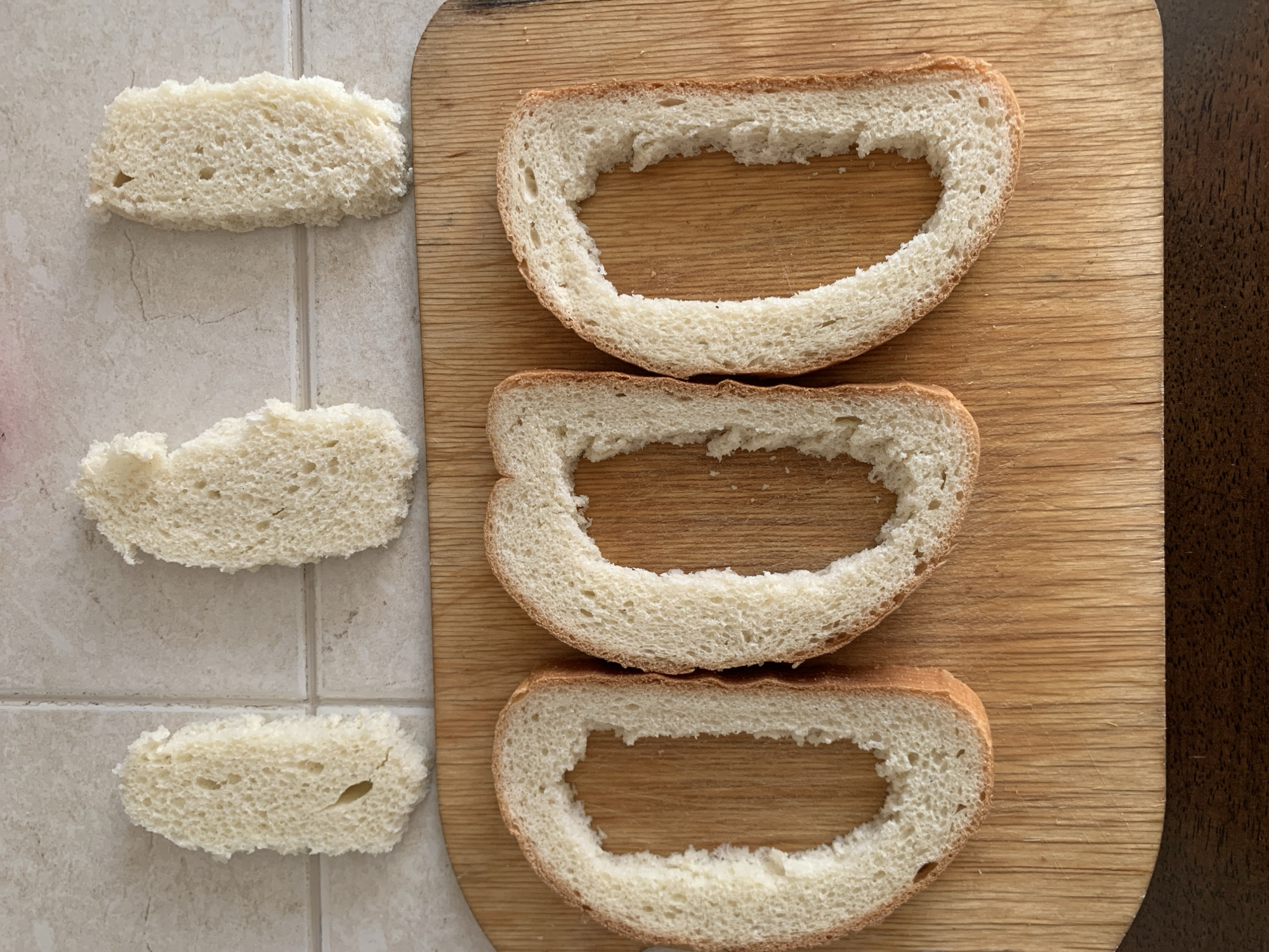 Three slices of bread, with the centers cut out, are arranged on a wooden cutting board. Above the cutting board, on a tiled surface, are three smaller pieces of bread, which appear to be the removed centers. The bread is light in color and has a porous texture. The cutting board is made of light-colored wood and shows visible grain. The tiled surface is a neutral, speckled color. The scene is lit from above, casting soft shadows. There are no people or animals present. The location is not identifiable as Kapyĺ, Belarus.