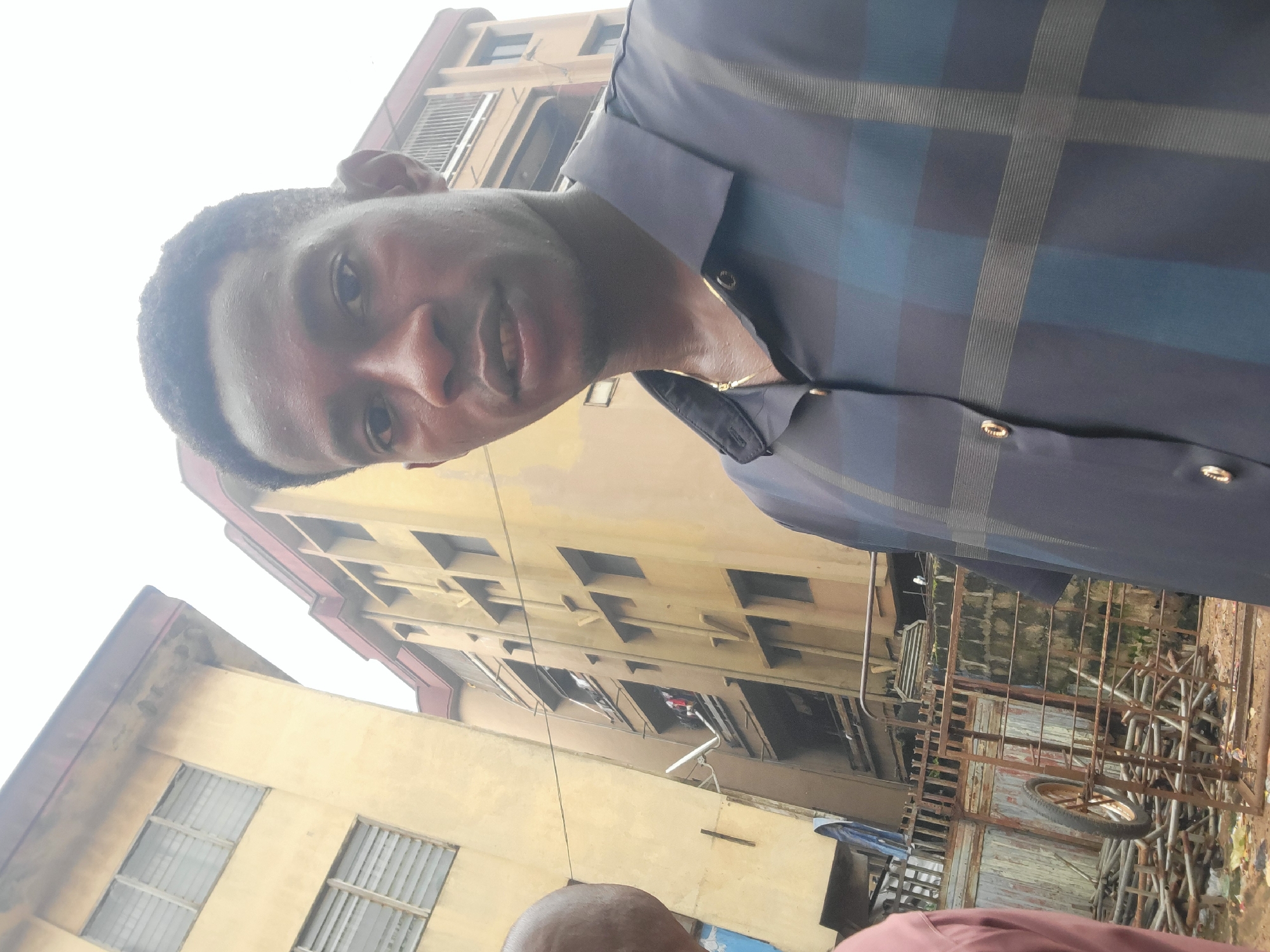 In Onitsha, Nigeria, a close-up selfie captures a young man in the foreground, his face damp with sweat, wearing a dark blue plaid collared shirt with gold buttons. He is positioned in front of a multi-story, weathered yellow building. Behind him, another similar building is visible, with balconies and windows that show glimpses of laundry hanging out. A rusted metal gate and a pile of construction materials, including a wheelbarrow, are in the lower left portion of the frame, suggesting an urban, possibly developing, environment. The sky is overcast, contributing to the muted lighting of the scene.