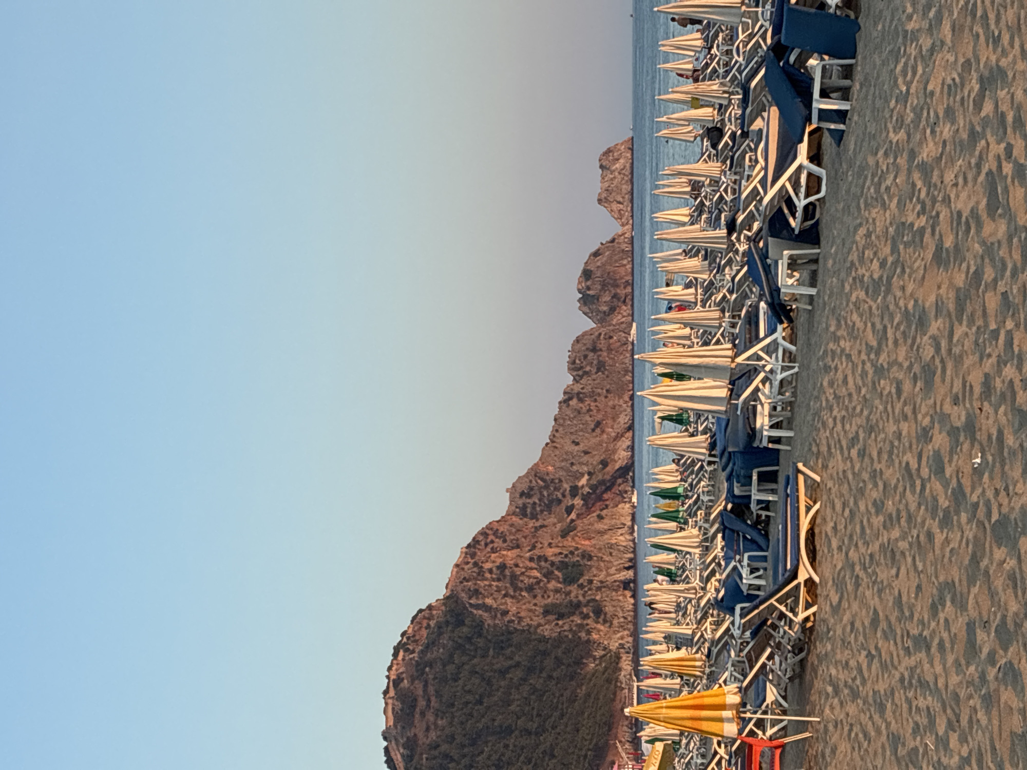 An outdoor, eye-level, full shot shows a beach in Alanya, Turkey, during the golden hour. The sand is in the foreground, with rows of lounge chairs and umbrellas stretching into the distance. A large mountain range dominates the background, with trees covering its slopes. The ocean is visible behind the beach, with a few boats sailing on its surface. The sky is clear and blue, with the sun casting a warm glow over the scene. The overall impression is one of a serene and peaceful beach, with the natural beauty of the landscape enhanced by the warm light of the setting sun. The beach is equipped with numerous lounge chairs and umbrellas, suggesting a popular tourist destination. The mountain is a prominent feature of the landscape, with its rugged terrain and dense vegetation adding to the natural beauty of the scene. The presence of boats on the water indicates that this is a coastal area, and the clear sky suggests good weather. The warm light of the setting sun creates a sense of tranquility and relaxation, making it an inviting place to spend time.