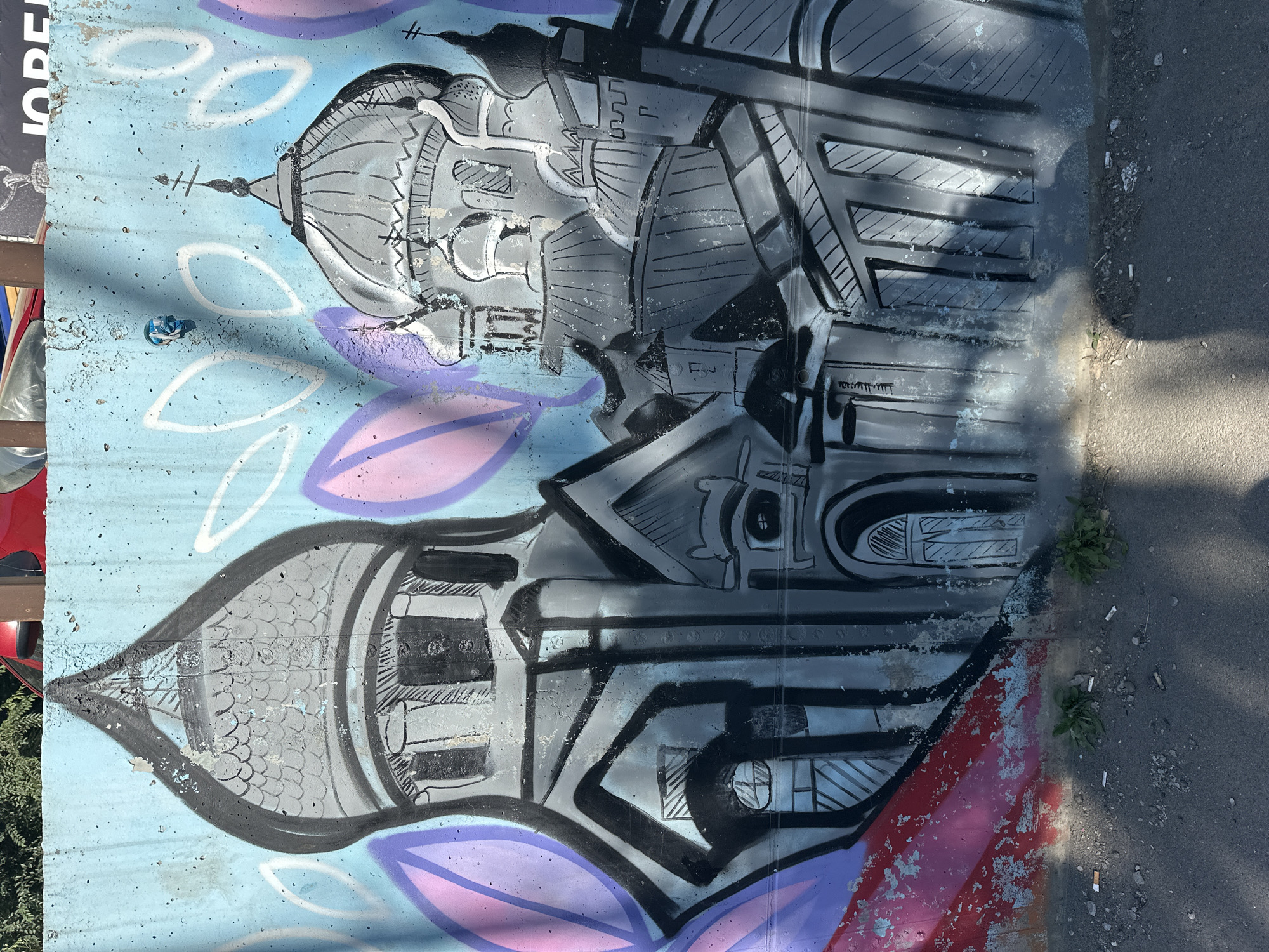The image shows a graffiti mural on a blue concrete wall, likely in the city of Biysk, Russia, given the context. The graffiti depicts a stylized rendition of several onion-domed buildings, resembling churches or cathedrals, rendered in black and gray paint. The buildings are depicted with architectural details like windows, arches, and cross-topped domes.
In the background, behind the graffiti, a red vehicle and a portion of another vehicle are visible at the top edge of the frame. The wall itself appears weathered and textured, with some peeling paint and visible concrete imperfections. The lower portion of the image shows the ground, a mixture of asphalt and dirt, with small patches of green grass and several discarded cigarette butts. Sunlight is casting shadows on the ground and the lower part of the wall, suggesting it is daytime. There are no people or animals present in the immediate scene. The overall impression is one of urban decay and street art.