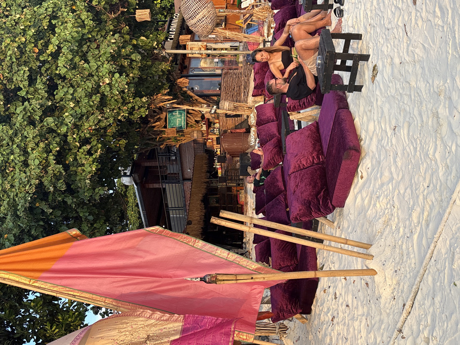 The image captures a beachside leisure area in Ban Ko Li Pe, Thailand, during golden hour. The foreground features fine white sand, marked with subtle undulations and footprints. On the left, tall bamboo poles support colorful fabric banners in shades of pink, orange, and gold. In the midground, multiple dark purple velvet or velour lounge chairs and ottomans are arranged directly on the sand.
A male and a female are seated on one of these lounge arrangements on the right. The male wears a black t-shirt, tan shorts, dark-rimmed glasses, and has a beard; he is looking down at a small rectangular object in his hands while holding a green bottle. The female, seated next to him, wears a black strapless top and has long dark hair, looking in his direction. Both are barefoot, with black sandals visible near the female's feet. A small, dark wooden square table stands in front of them. To their left, a second male is visible, lying back on a purple lounge with his feet elevated.
The background reveals a two-story establishment with a dark wooden and brick facade, partially covered by a thatched roof. A large, densely leafed green tree dominates the center background, with various woven decorations or lamps hanging from its branches and the building structure. Signs visible on or near the building include "CHARMINGO" and "HAPPY VIBE," suggesting a bar or restaurant setting with bottles lining a counter. The overall scene depicts a relaxed environment at a beachfront venue.