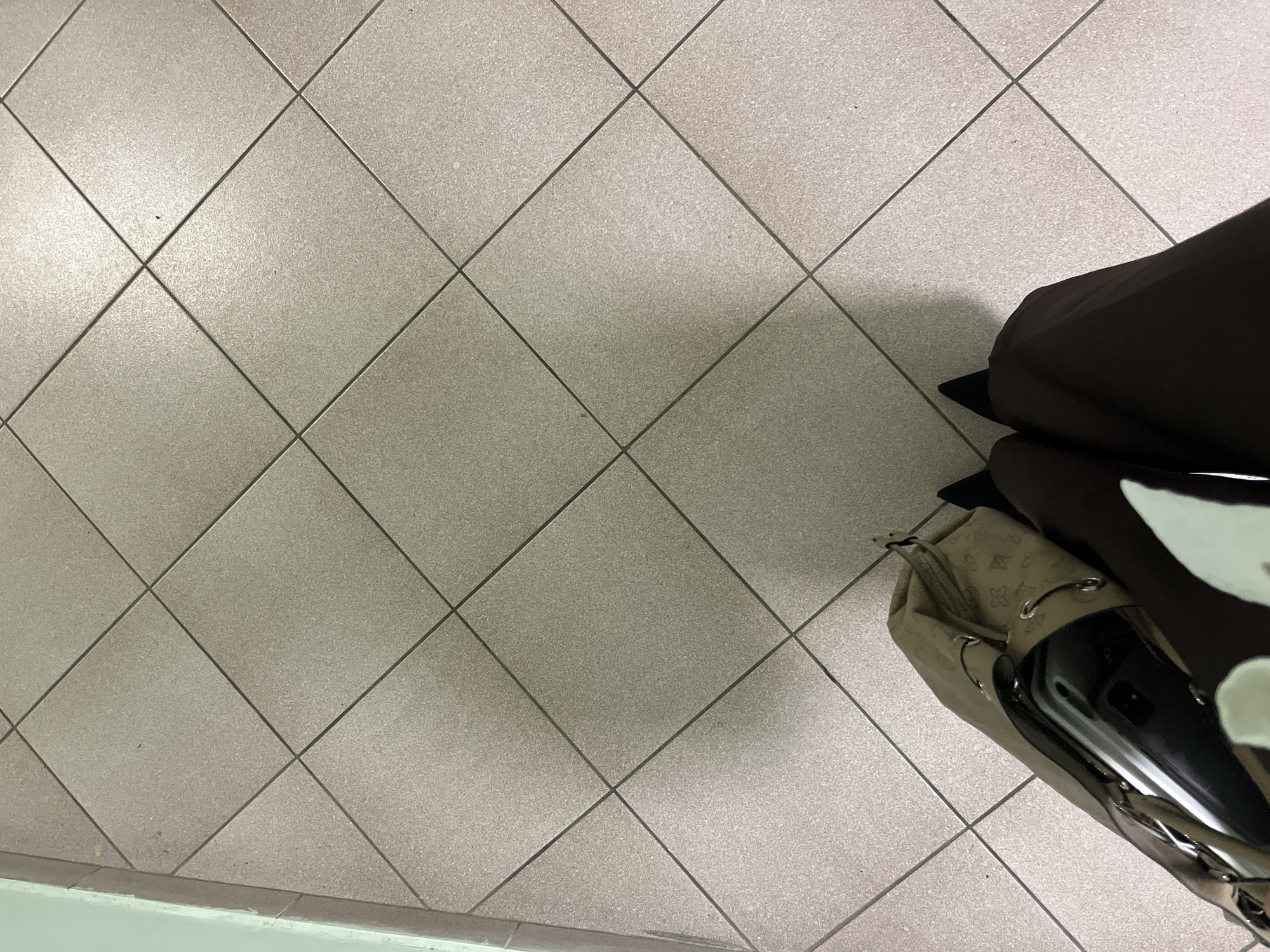 A tiled floor in Moscow, Russia, features gray square tiles arranged in a diamond pattern. In the lower right corner, a brown bag and a lighter-colored purse with straps rest on the tiles. Part of a black object is visible at the top. The tiles are clean with a subtle texture. Along the bottom left, a white baseboard is present.