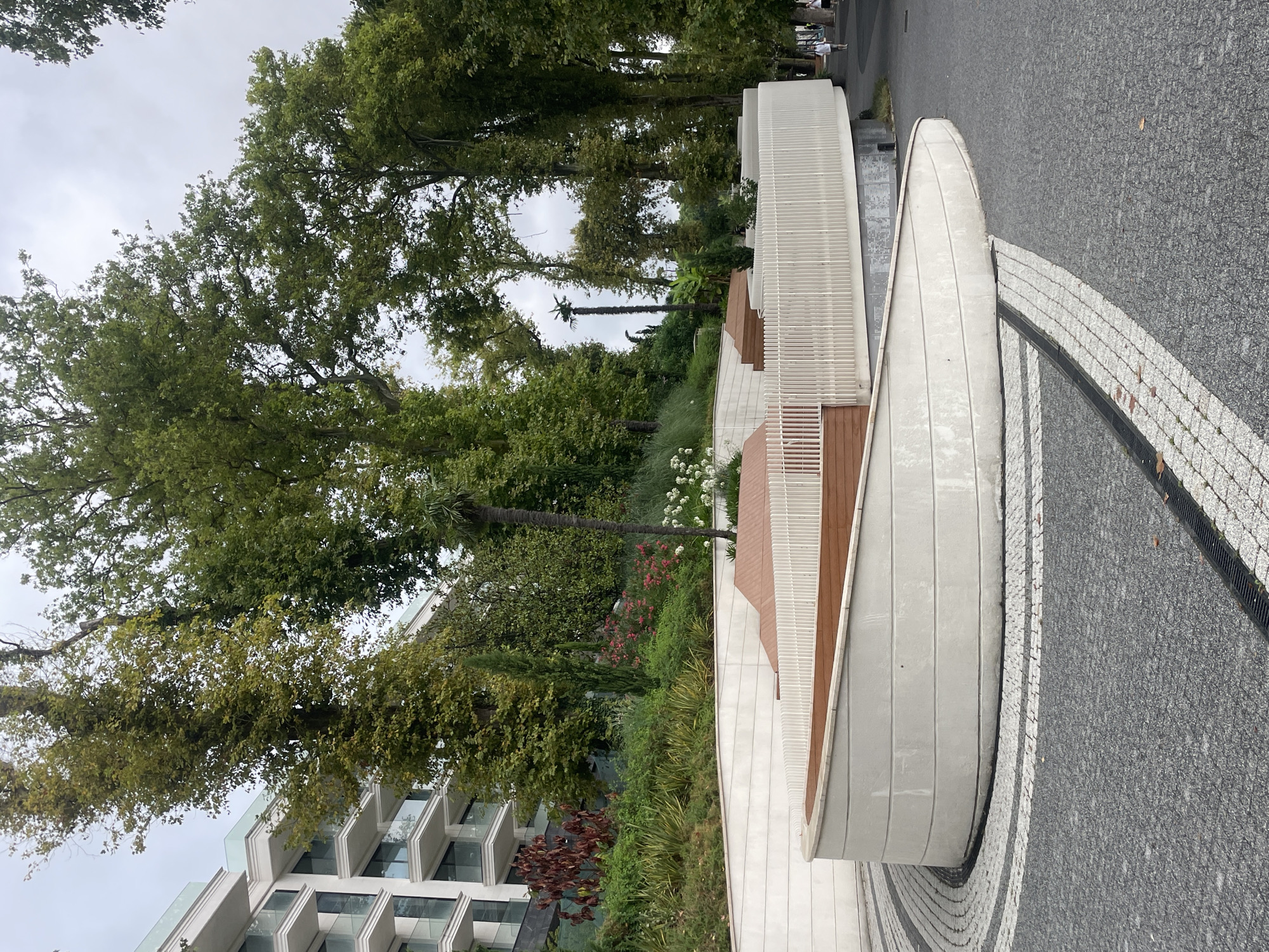 This image shows a modern, curved, concrete structure with wooden seating and a white picket fence. The structure is surrounded by a patterned cobblestone walkway and a darker asphalt path. Behind the structure, there is lush greenery, including tall trees, bushes, and flowering plants. A modern white building with multiple balconies is visible in the background. The scene appears to be in a park or public space. The location is the Sochi Arboretum in Sochi, Russia. A few pedestrians are visible walking on the asphalt path in the distance.