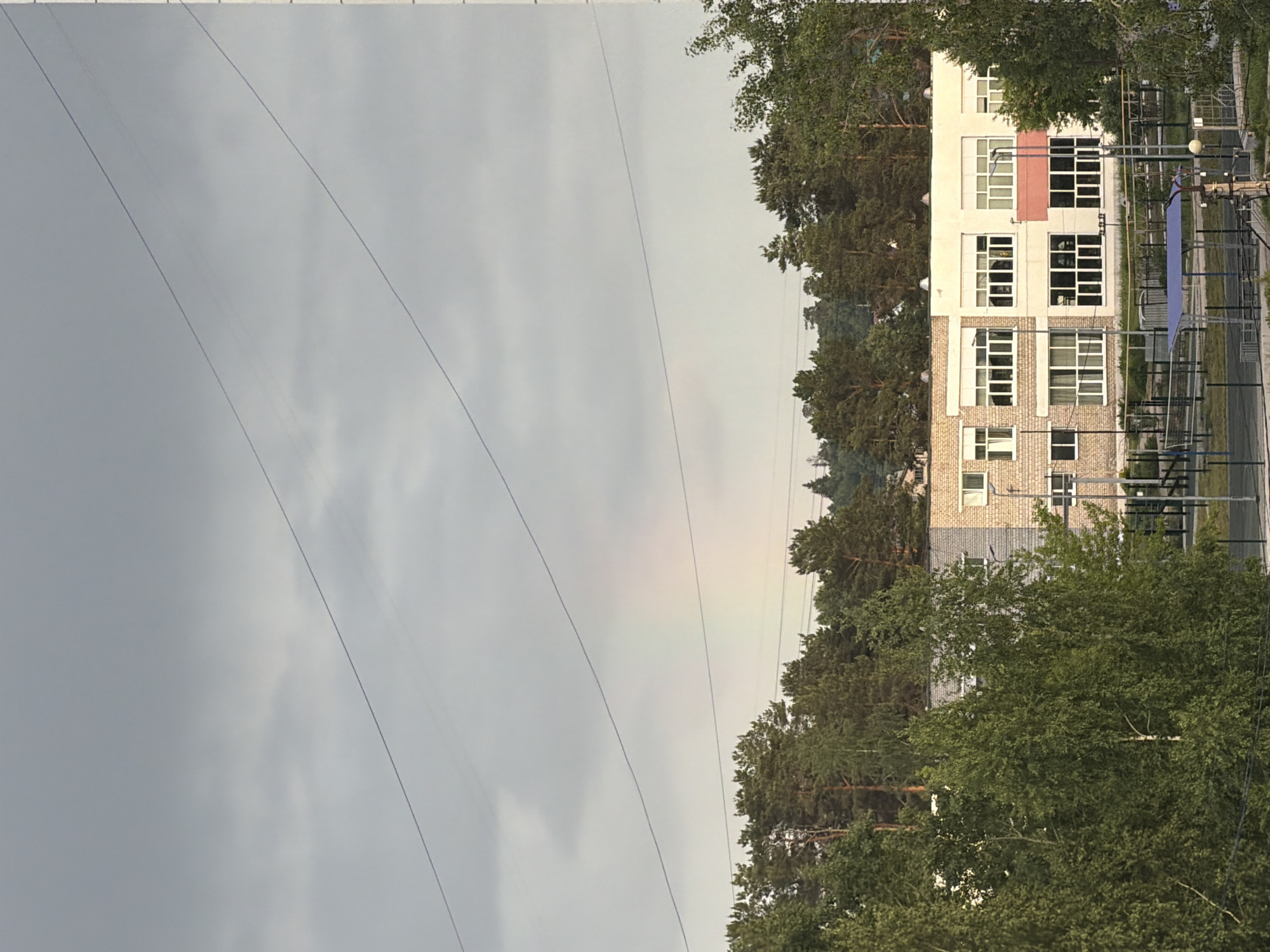 The image depicts a scene in Chaikovsky, Russia, showing a residential area with a view of a light-colored building partially obscured by trees. Power lines cut across the grey sky on the left side of the frame. A portion of a rainbow is faintly visible in the sky near the trees. A playground or recreational area is partially visible next to the building, with a fence and some structures.