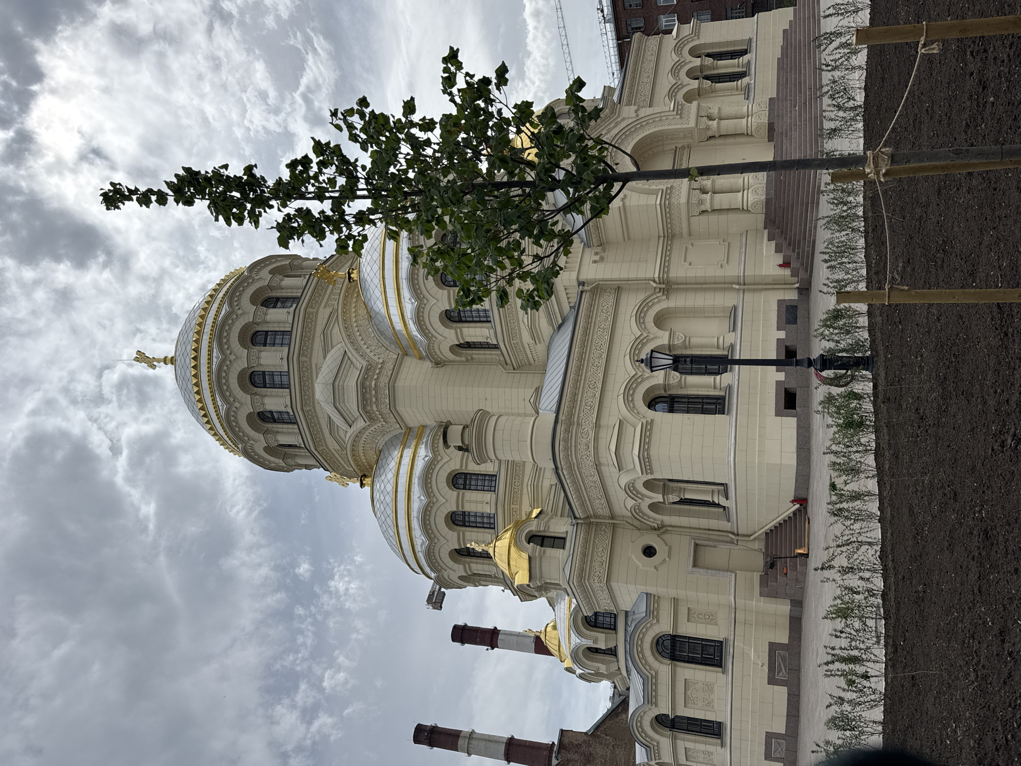 The image shows the Naval Cathedral of Saint Nicholas in Kronstadt, a city within the federal city of Saint Petersburg, Russia. The grand, cream-colored cathedral features multiple golden domes topped with crosses. Its facade is ornate with arched windows and detailed architectural elements. In the foreground, a young tree is planted, supported by wooden stakes and rope. Behind the cathedral, two industrial chimneys with red and white banding are visible. The sky is overcast with dramatic clouds. The ground in front of the cathedral is paved, with a small landscaped area featuring young bushes. Stairs lead up to the cathedral's entrance.