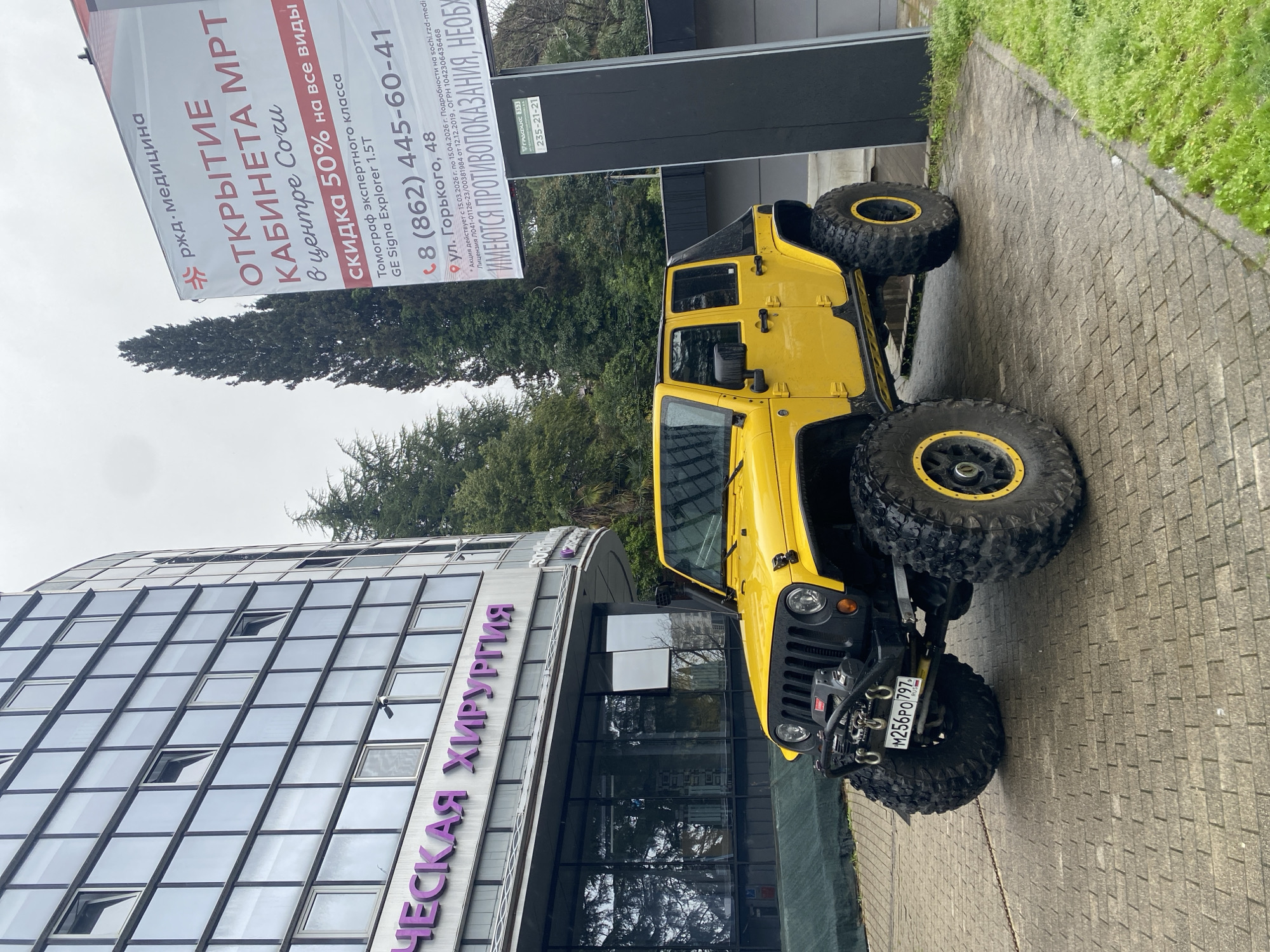 The image shows a bright yellow four-door Jeep Wrangler parked on a paved sidewalk next to a building. The building's facade is predominantly glass, and its name "ЧЕСКАЯ ХИРУРГИЯ" is displayed in purple lettering. The Jeep has large, off-road tires with yellow rims. A license plate, "M256PO 797," is visible on the front of the vehicle.
Behind the Jeep and building, there's a billboard ad for "РЖД Медицина" promoting the opening of an MRI facility in the center of Sochi, offering a 50% discount. The advertisement lists the address as "ул. Горького, 48" (Gorky Street, 48), and a phone number. Trees are visible behind the billboard. The environment appears urban and slightly overcast.