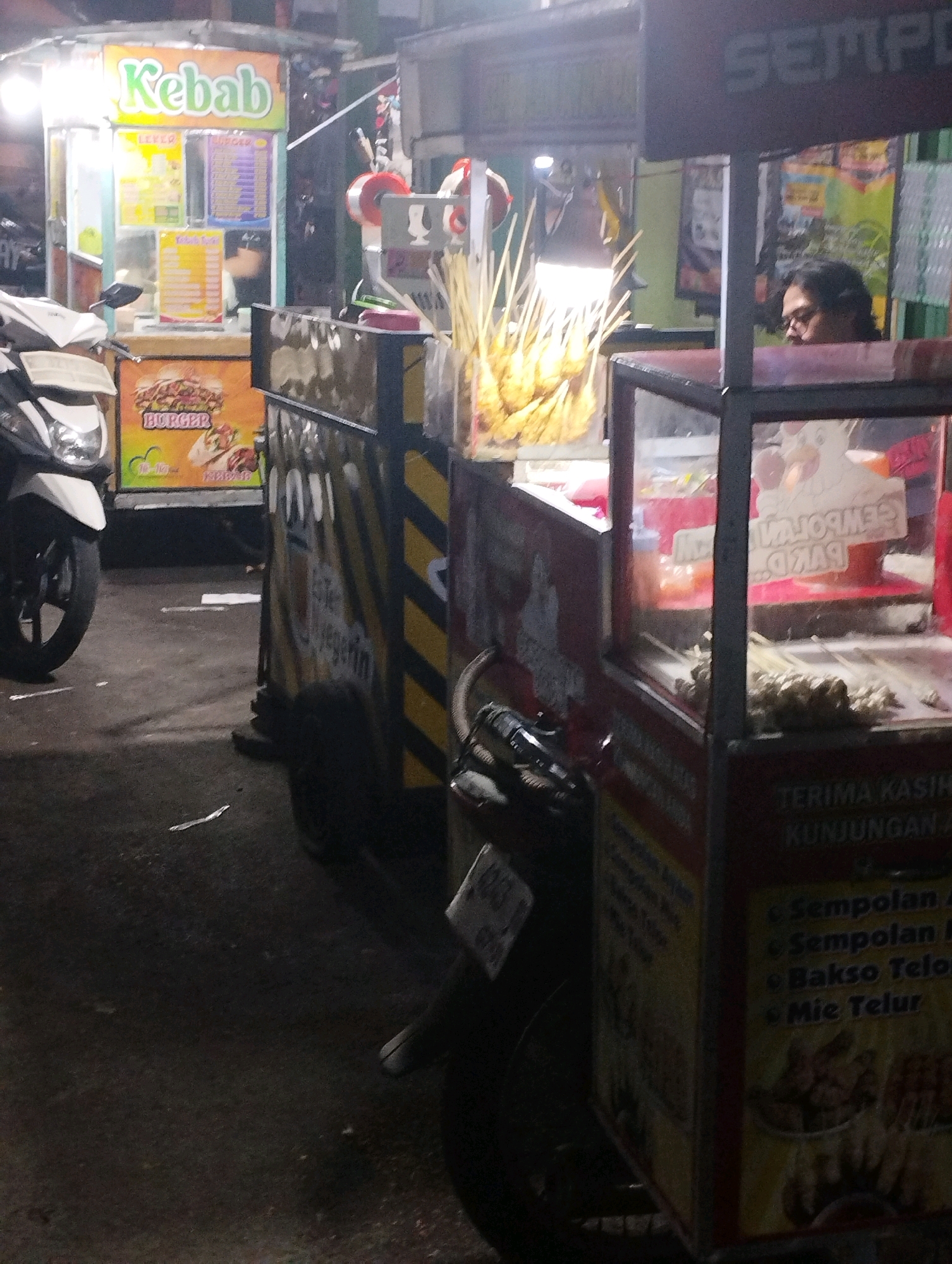 A dimly lit, nighttime street scene in Kecamatan Pati, Indonesia, features two food stalls and a motorcycle. To the left, a "Kebab" food stall is brightly illuminated, displaying menus and food items. In front of it, a white motorcycle is parked. To the right, a second food stall, identified by the partially visible "SEMPO," is less illuminated. A person with dark hair and glasses is visible behind the counter of this stall. Skewers of food are displayed prominently in both stalls. The ground is dark and appears to be asphalt, with scattered debris. The overall impression is that of a typical street food vendor scene at night.