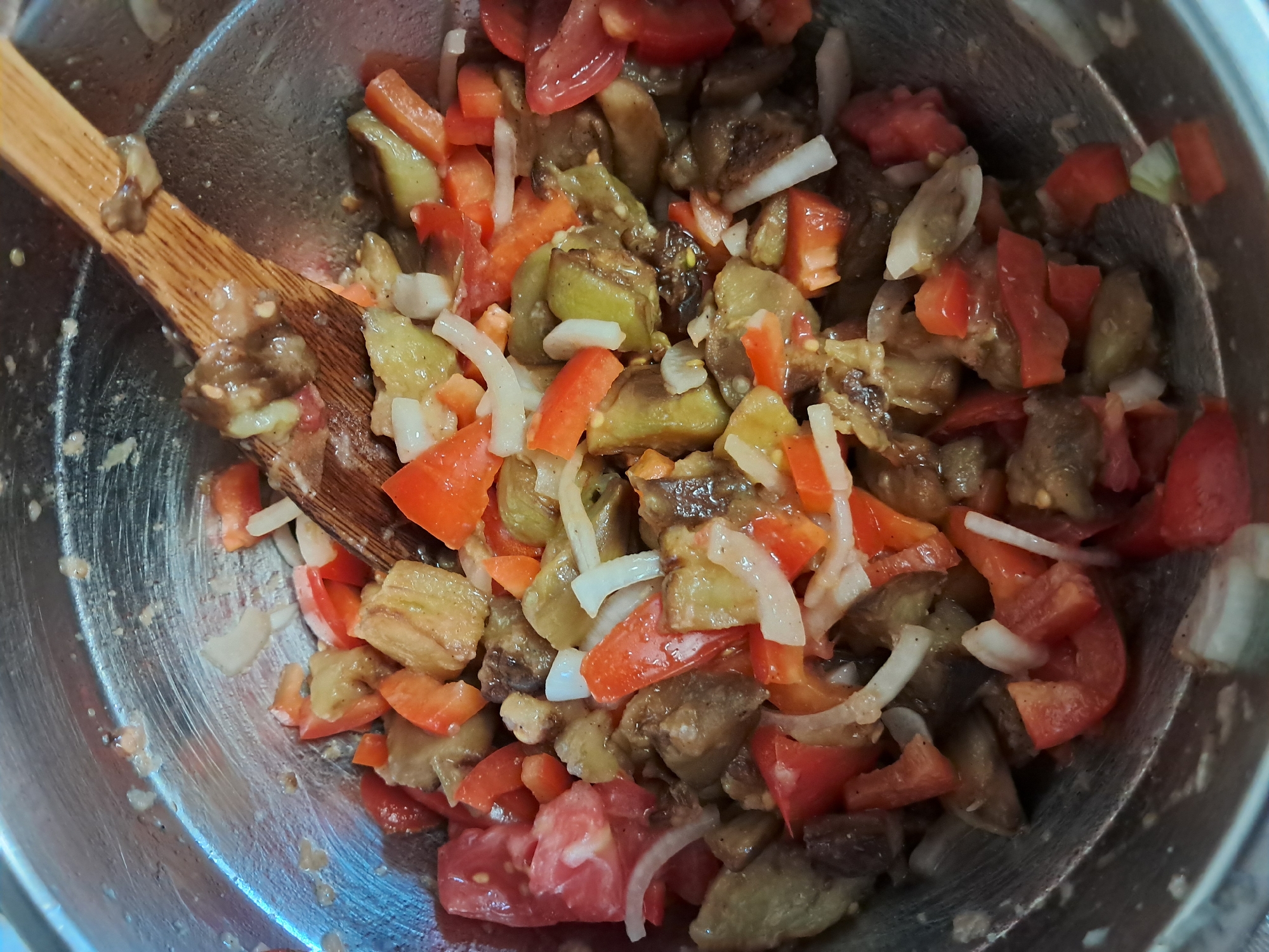 A close-up, overhead shot shows a metal bowl filled with a chopped mixture of cooked eggplant, tomatoes, and onions. A wooden spoon is partially submerged in the mixture, appearing to stir or serve it. The ingredients are glistening, suggesting they are moist or have a dressing. The bowl is metallic and has a brushed texture, with some remnants of the food clinging to the sides. The overall scene is one of food preparation. The image appears to be taken indoors, with no discernible urban environment details.