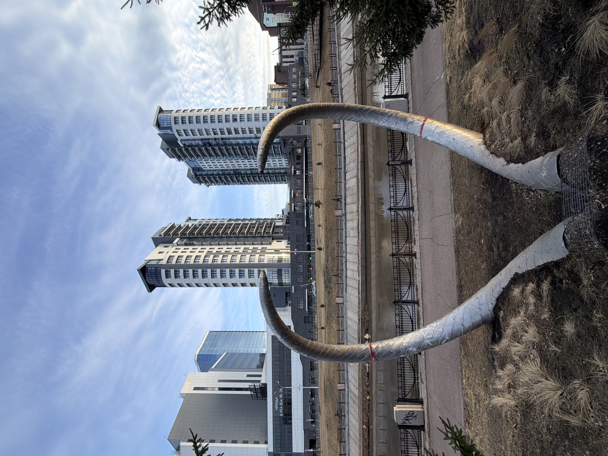 The image shows a riverside scene in Krasnoyarsk, Russia. In the foreground is a large, silver, stylized sculpture resembling a pair of mammoth tusks. The sculpture is located on a grassy area next to a paved walkway and a metal fence bordering a river or canal. Behind the sculpture are several modern high-rise buildings with light-colored facades and numerous windows. Other buildings of varying heights and architectural styles are visible further back, suggesting an urban environment. The sky is partly cloudy, with patches of blue visible. Vegetation, including grass and trees, can be seen along the riverbank. The scene appears to be taken during the daytime, likely in the spring or fall.