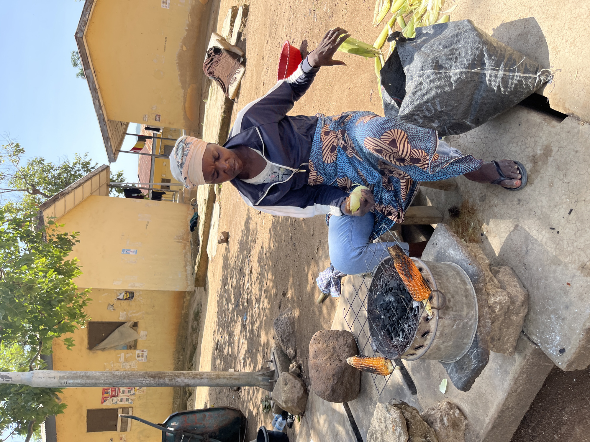 In a sunlit outdoor setting in Rafin Par, Nigeria, a woman is preparing and roasting corn. She is seated on a low wooden stool, dressed in a light-colored headwrap, a dark blue and white jacket, a patterned blue and brown skirt over light blue jeans, and flip-flops. She holds a partially husked ear of corn in her left hand, her right hand reaching towards a pile of unhusked corn beside her.
An improvised grilling station is positioned in front of her, consisting of a metal drum containing charcoal, topped with a metal grid. Two ears of corn are actively roasting on this grill, supported by concrete blocks. Near the woman, a large black woven sack, partially labeled "FUL," holds more corn, and a red plastic bowl is visible.
The background features several single-story buildings with light yellow-ochre walls and simple roofs, indicative of a rural or semi-urban settlement. Faded posters and some text, including "B.A. OF P." on one wall, are affixed to the building surfaces. The ground is bare earth, and green foliage from a tree is visible on the left. The scene is brightly lit by the sun, suggesting it is midday or early afternoon under a clear sky. The woman appears focused on her task.
