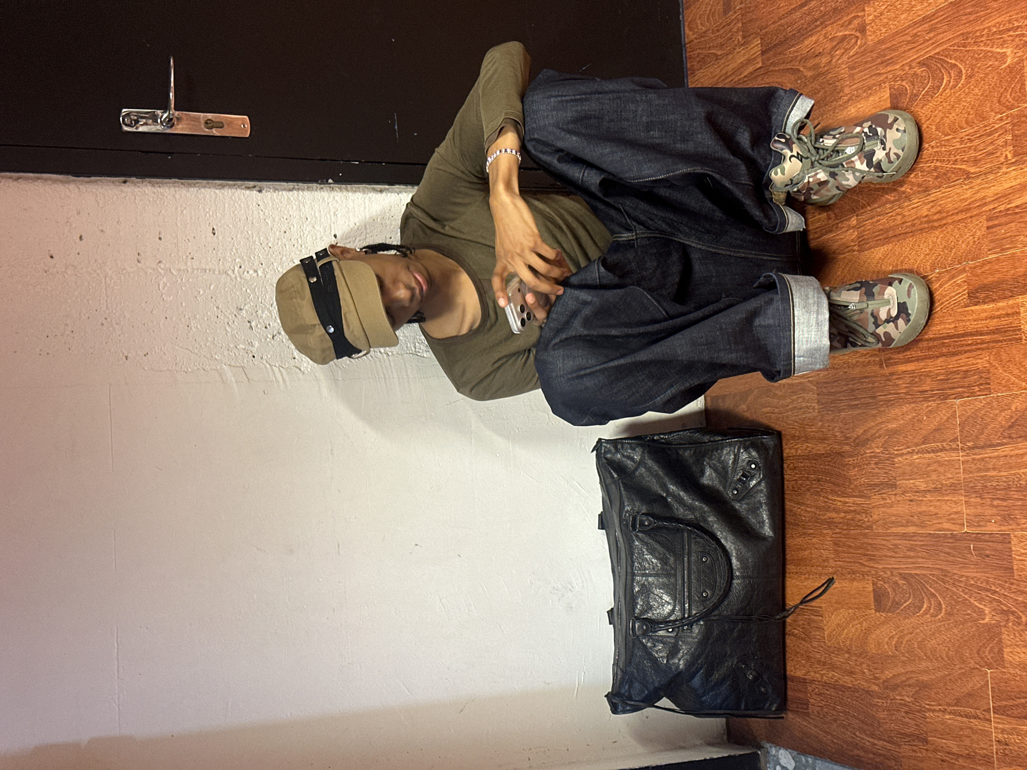 A person wearing a tan hat, a long-sleeved olive shirt, dark denim jeans, and camouflage shoes is seated on the floor. They are holding a mobile phone. Beside the person is a black leather duffel bag. The person is leaning against a white wall with a dark door behind them. The floor is wood. The location is unclear, and cannot be determined to be in Paris, France.