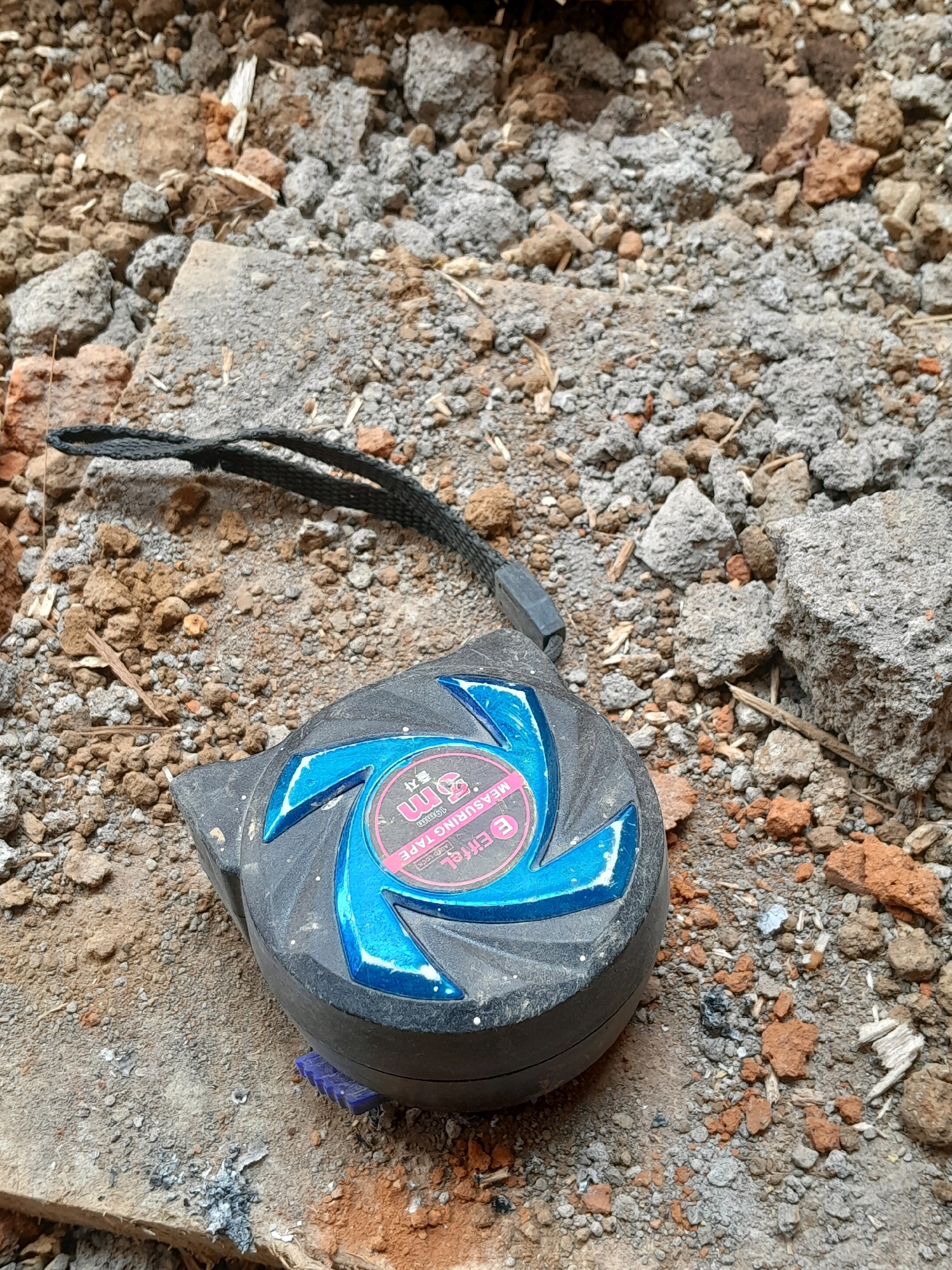 A black tape measure with a blue, swirled decorative pattern lies on a surface of dirt, small rocks, and broken concrete. A black strap is attached to the tape measure. The scene is outdoors, with the ground showing signs of construction or demolition debris. The tape measure has a label that includes "3m" and "MEASURING TAPE". The overall impression is of a construction site or area undergoing renovation. This image was taken in Karangduren, Indonesia.