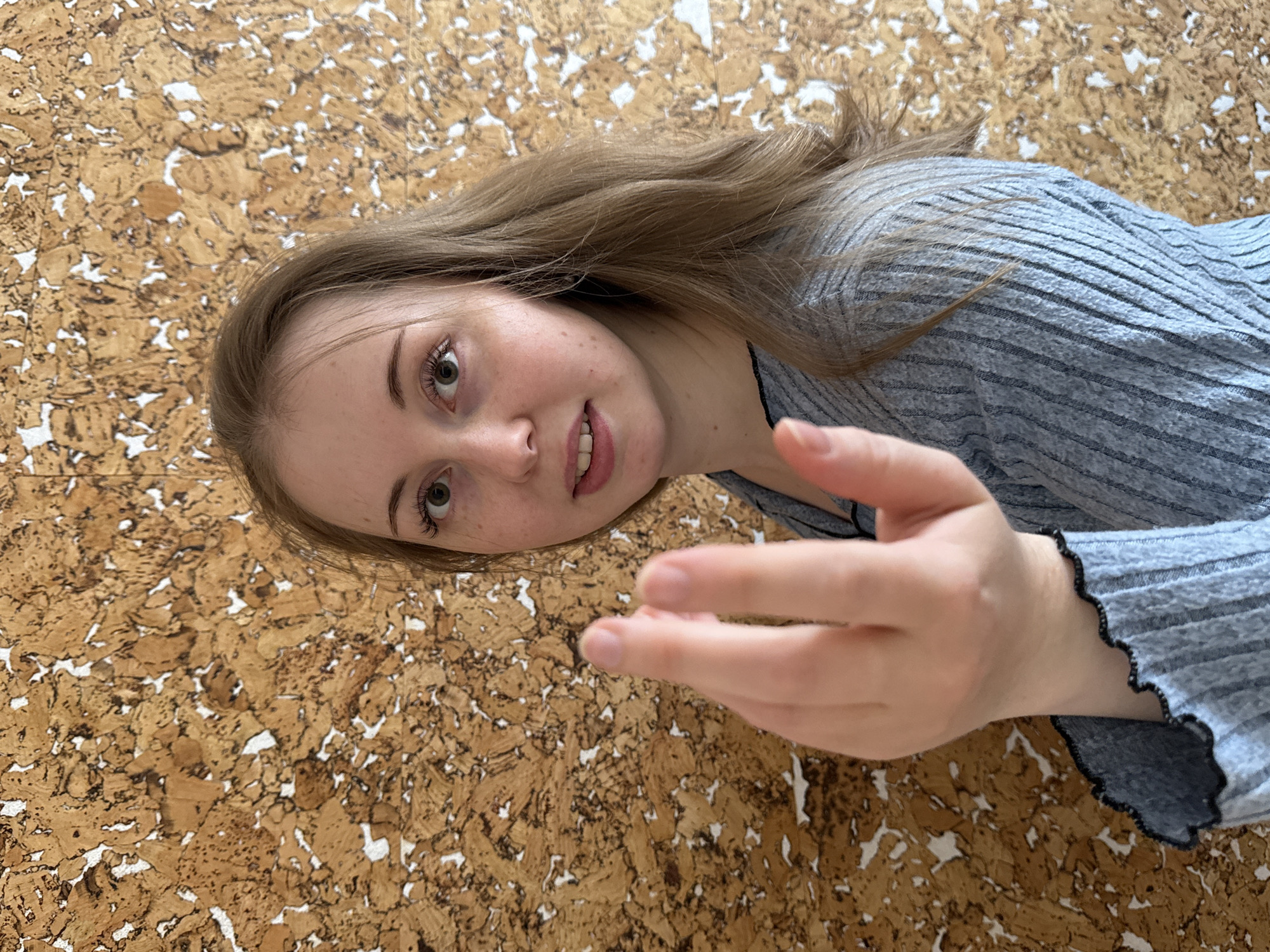 An image captures a young woman from a slightly elevated perspective, with the frame rotated clockwise. She has long, light brown hair and fair skin, with visible freckles across her nose and cheeks. Her eyes are a light greenish-brown, and she has naturally arched eyebrows. Her mouth is slightly open in a gentle smile, revealing some teeth. She is wearing a light blue or grey ribbed long-sleeved top, which features a dark, ruffled or scalloped trim on the cuffs.
The woman is positioned on a textured surface that appears to be made of cork or a similar granular material, characterized by irregular brown shapes scattered over a white background. Her head is tilted, and she is looking upwards, directly towards the viewer. Her right hand is extended into the foreground, fingers slightly splayed, as if reaching out or making a gesture. The overall impression is one of direct engagement with the camera. The setting appears to be indoors with soft, even lighting. There is no visible text in the image.