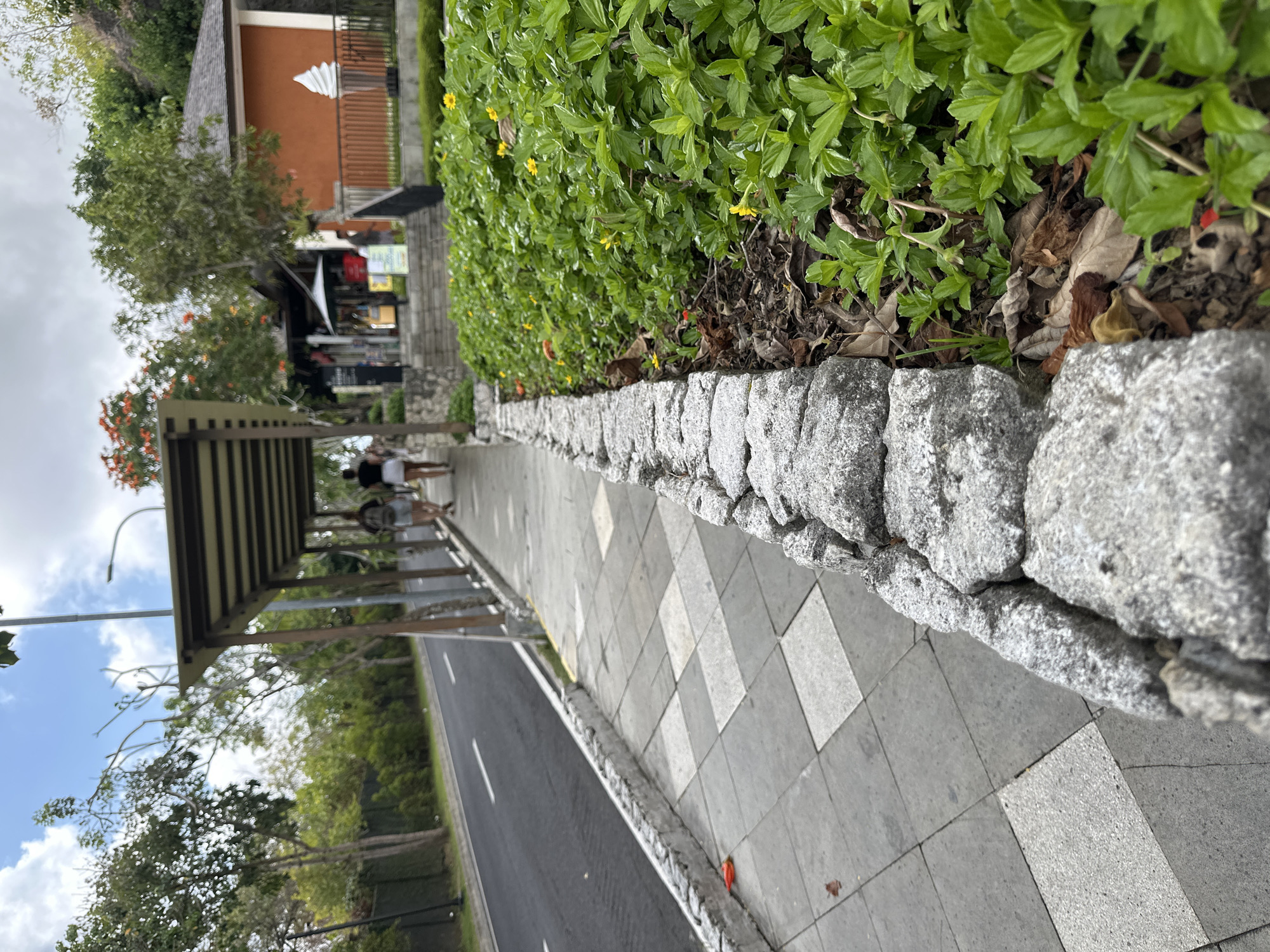 The image depicts a sidewalk in Kuta Selatan, Indonesia, next to a road. Two women are walking away from the camera on the sidewalk, which is covered by a shaded canopy. The sidewalk is paved with square tiles, and a stone wall borders a lush green garden on the right. The garden is filled with small yellow flowers and some orange blossoms. In the background, there's an orange building with a white ice cream cone sculpture in front of it, suggesting a commercial establishment like an ice cream shop or cafe. The sky is cloudy with patches of blue. The overall scene suggests a warm, tropical environment.