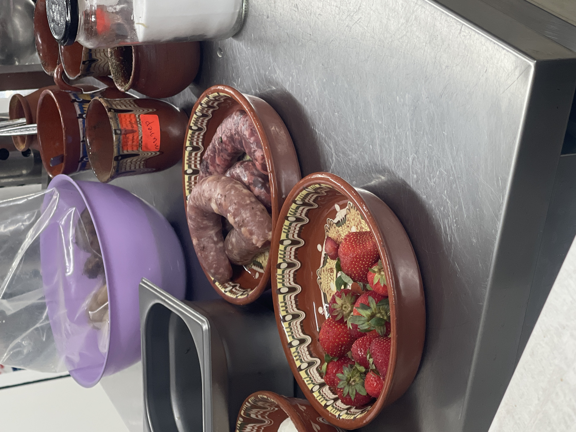 The image displays a close-up, slightly overhead view of various food items and kitchenware arranged on a metallic counter. No individuals are visible.
In the foreground, a brown ceramic bowl with decorative white, yellow, and black patterns contains a mound of fresh, red strawberries. To its left, another similar ceramic bowl holds several coiled, raw sausages. A silver metal counter, heavily scratched, serves as the primary surface. Further to the left, a purple plastic bowl is partially visible, next to a silver metal insert or container.
Towards the upper left, several small, brown ceramic mugs or pots are arranged. One clear glass jar with a black lid, possibly containing salt or sugar, rests among them. One ceramic mug visibly contains dried green herbs, while another features a distinctive folk art pattern and has a red sticker affixed to its side. The text on this red sticker appears to be in Cyrillic script, reading "мuнeр" or a similar word, which might denote a type of herb, spice, or a brand.
The scene suggests a food preparation area, possibly a kitchen, with ingredients laid out. The traditional ceramic pottery hints at a cultural aesthetic, potentially Eastern European. The time of day and weather are not discernible from the indoor setting.