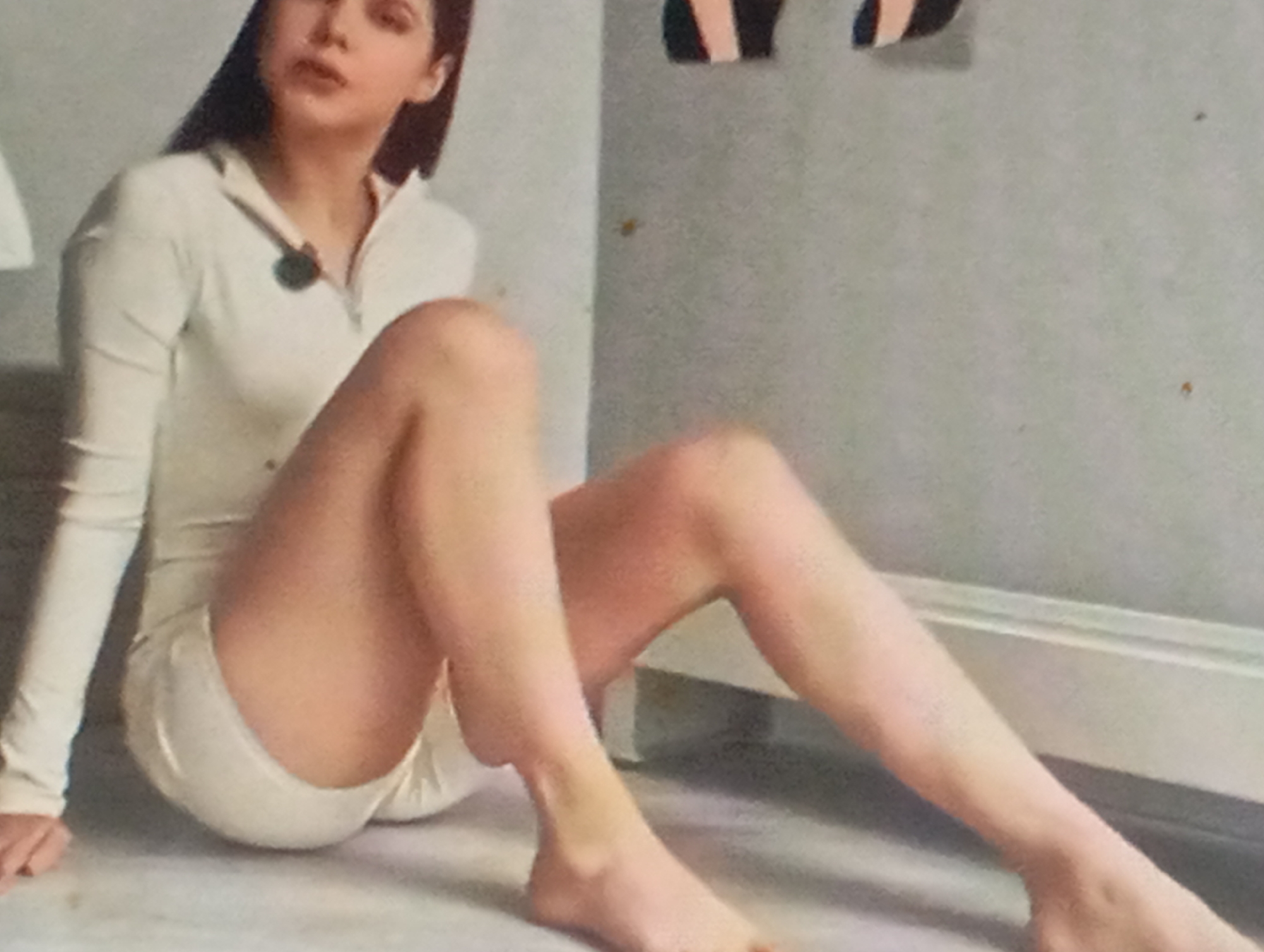 The media file shows a medium shot of a young woman sitting on the floor, leaning against a grey wall. She has long, dark hair and is wearing a white, form-fitting outfit that appears to be a jumpsuit or a romper with long sleeves. A dark pendant hangs from her neck. Her legs are crossed, and her right leg is extended forward. The setting appears to be indoors, with a simple, light-colored floor and a white baseboard along the wall. Above and behind her, a portion of some black and white striped objects is visible on the wall, possibly artwork or decoration. The lighting is soft, suggesting it could be daytime indoors or artificial lighting. The woman's expression is neutral, and her gaze is directed slightly away from the camera. There are a few small specks on the wall, possibly dust or marks. The overall impression is a posed, casual shot.