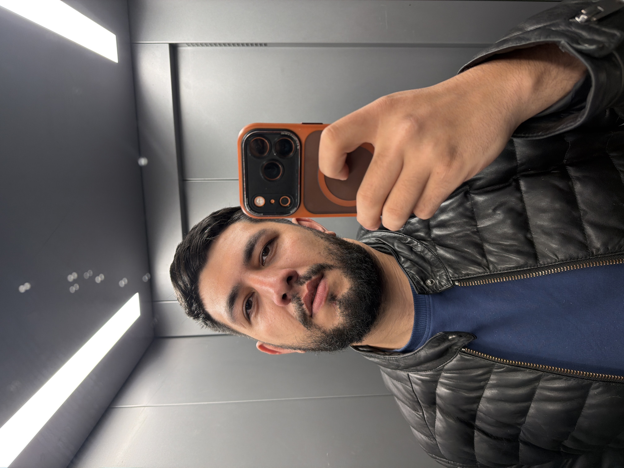 A selfie taken by a man with dark hair and a beard, wearing a blue shirt and a black leather jacket inside an elevator with gray walls and a long horizontal light fixture on the ceiling. The man is holding an orange and brown phone with multiple lenses.