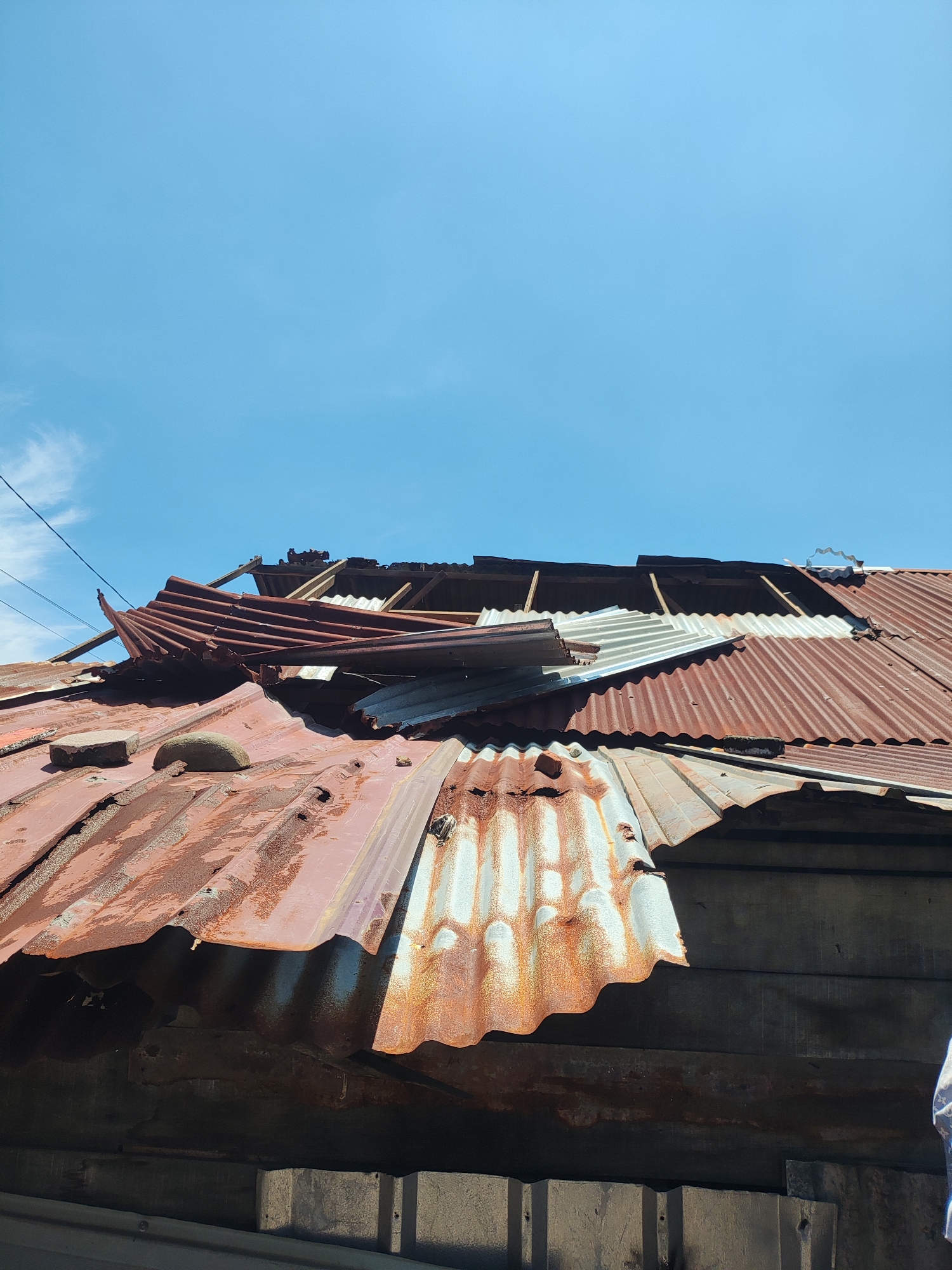 The image shows a dilapidated roof of a building in Lhokseumawe, Indonesia. The roof is made of corrugated metal sheets, heavily rusted and partially collapsed. There are several large holes exposing the wooden frame underneath. A few pieces of the roofing material are dislodged and hanging precariously. Two rocks sit atop the corrugated metal on the left side of the image. The background is a bright blue sky with sparse clouds.