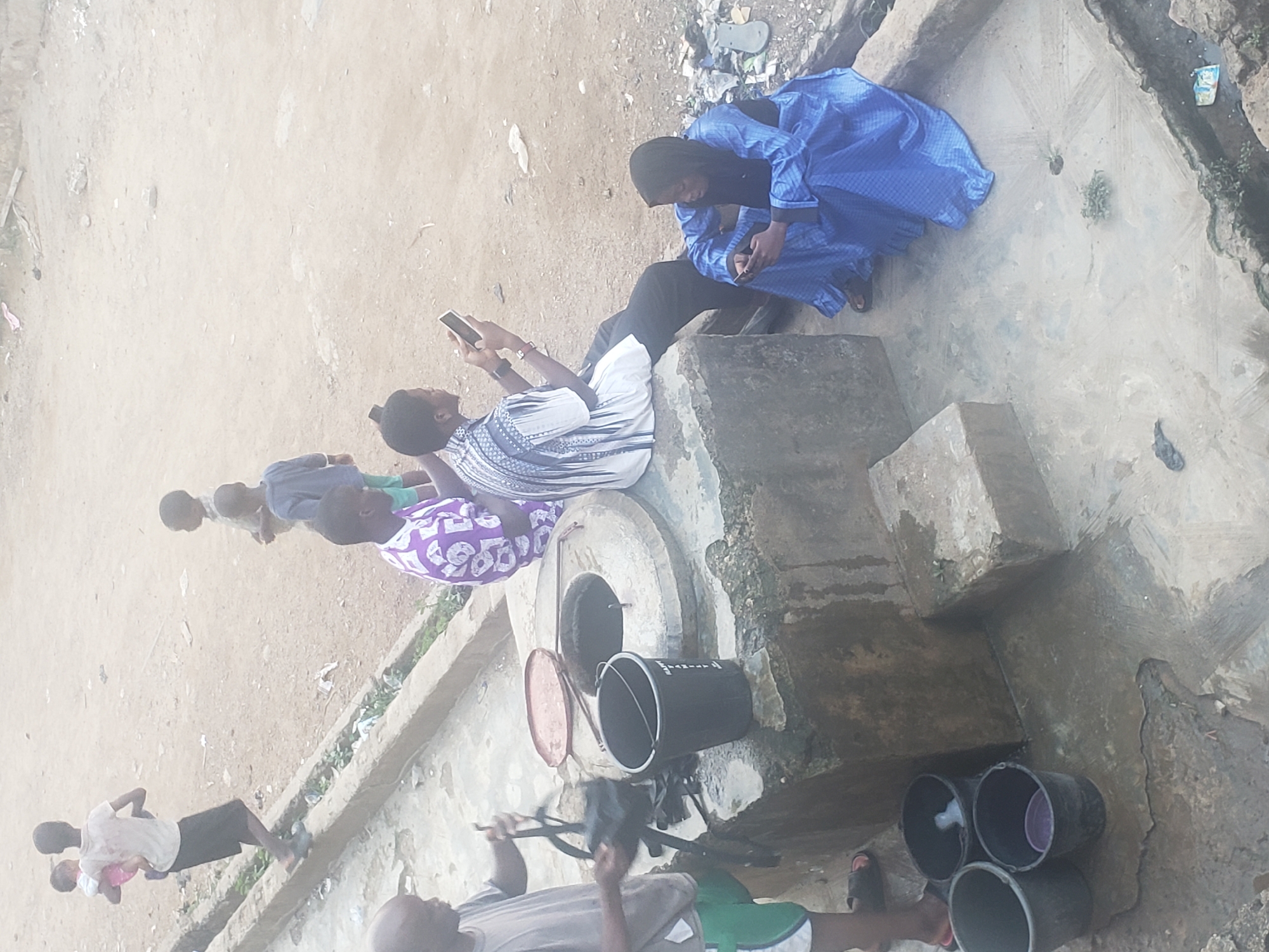 The image, taken from an elevated perspective, depicts a scene in Alalubosa, Nigeria.
People: Several individuals are present. Two women are seated on a concrete structure, each engrossed in their respective mobile phones. Nearby, other people are walking. One man is holding a child, while another person appears to be lifting a water bucket.
Objects: A well is visible with multiple buckets, some stacked, others positioned to draw water. One bucket has a lid placed to its side. The women are seated on concrete structures, and mobile phones are held by both women.
Actions: People are seen engaging in various activities, including using mobile phones, collecting water from a well, walking, and holding a child.
Interactions: There is no visible interaction between the women using their mobile phones, but other people appear to be engaged in conversation.
Environment: The location appears to be an outdoor, communal area, possibly a residential neighborhood with a well. The ground is dusty. The overall ambiance suggests a somewhat humid climate.