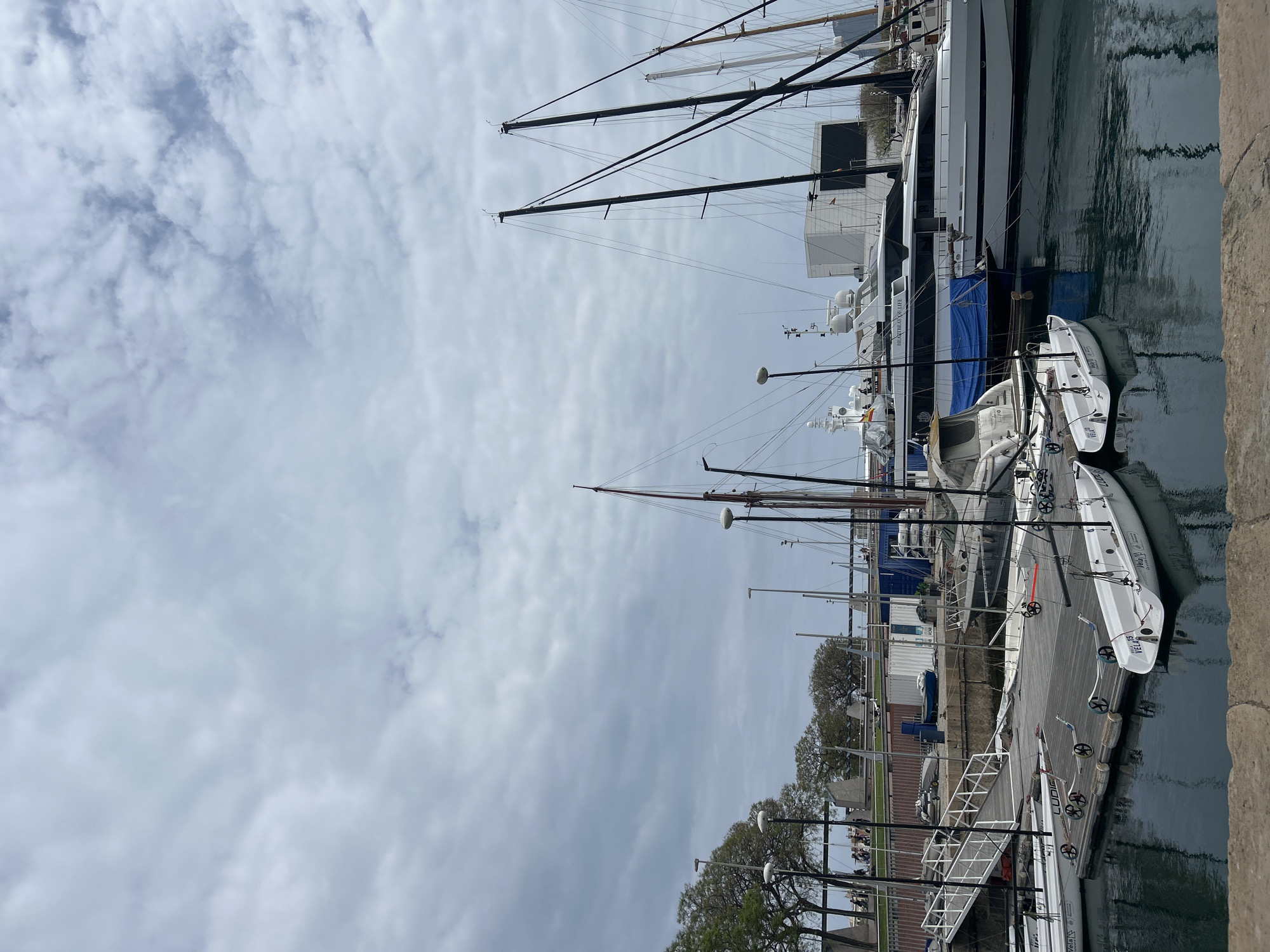 The image shows a marina scene on an overcast day, likely located in Barcelona, Spain. Several large yachts are docked, distinguished by their tall masts and sophisticated superstructures. Smaller white boats, possibly dinghies or tenders, are tied alongside the docks and larger vessels. Dockside structures and trees line the water's edge, obscuring any distinct city landmarks. The sky is covered with layered gray clouds.