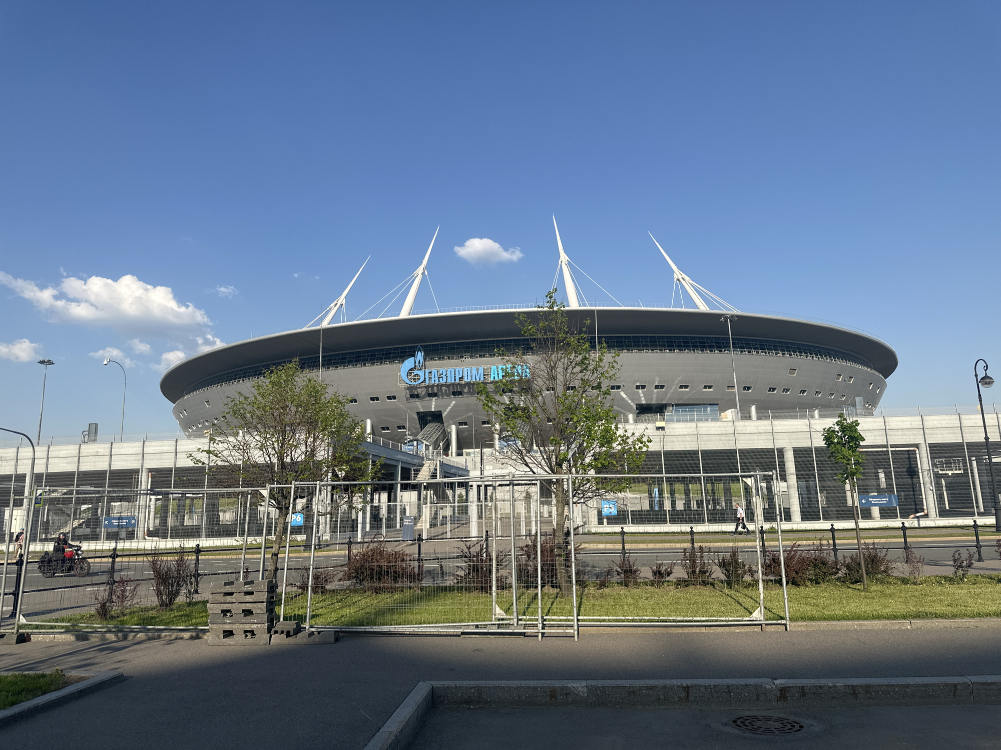 The image shows the Gazprom Arena stadium in Saint Petersburg, Russia, on a sunny day with a blue sky and a few white clouds. The stadium is a large, oval-shaped structure with a gray exterior and a white roof. Four tall, white pylons protrude from the roof. The words "GAZPROM ARENA" are displayed in blue lettering on the side of the stadium. A metal fence runs along the perimeter of the stadium, with some grass and bushes in front of it. There is a street in the foreground with a sidewalk and a manhole cover. A motorcyclist is riding on the street, and a pedestrian is walking on the sidewalk. Two young trees grow on the green space between the fence and the road. A streetlamp is visible on the right side of the image.