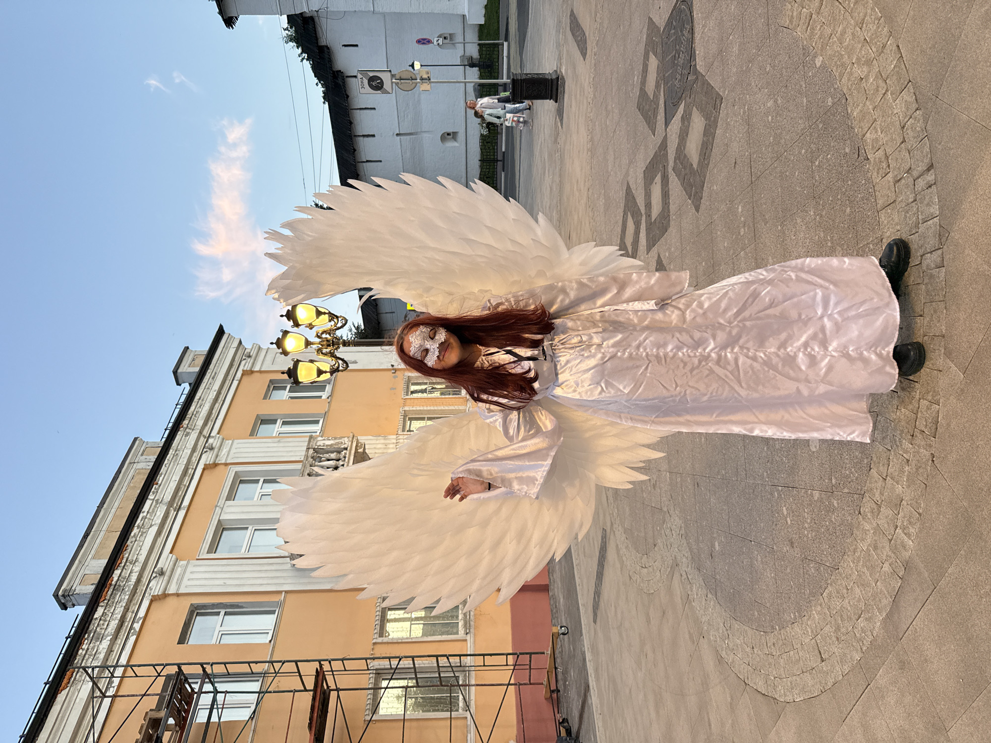 A young woman wearing a white satin robe, large white feathered wings, and a white lace mask stands in a paved plaza in Zhukovo, Russia. She has long auburn hair and is looking towards the camera. In the background, there is a light orange building with scaffolding on the left side, and a white building with a dark roof on the right. A lamppost with three lights is positioned behind the woman. The sky is a light blue with a few wispy clouds. The ground is paved with stones and there are some diamond-shaped patterns. Two people are walking in the background on the right side of the image.