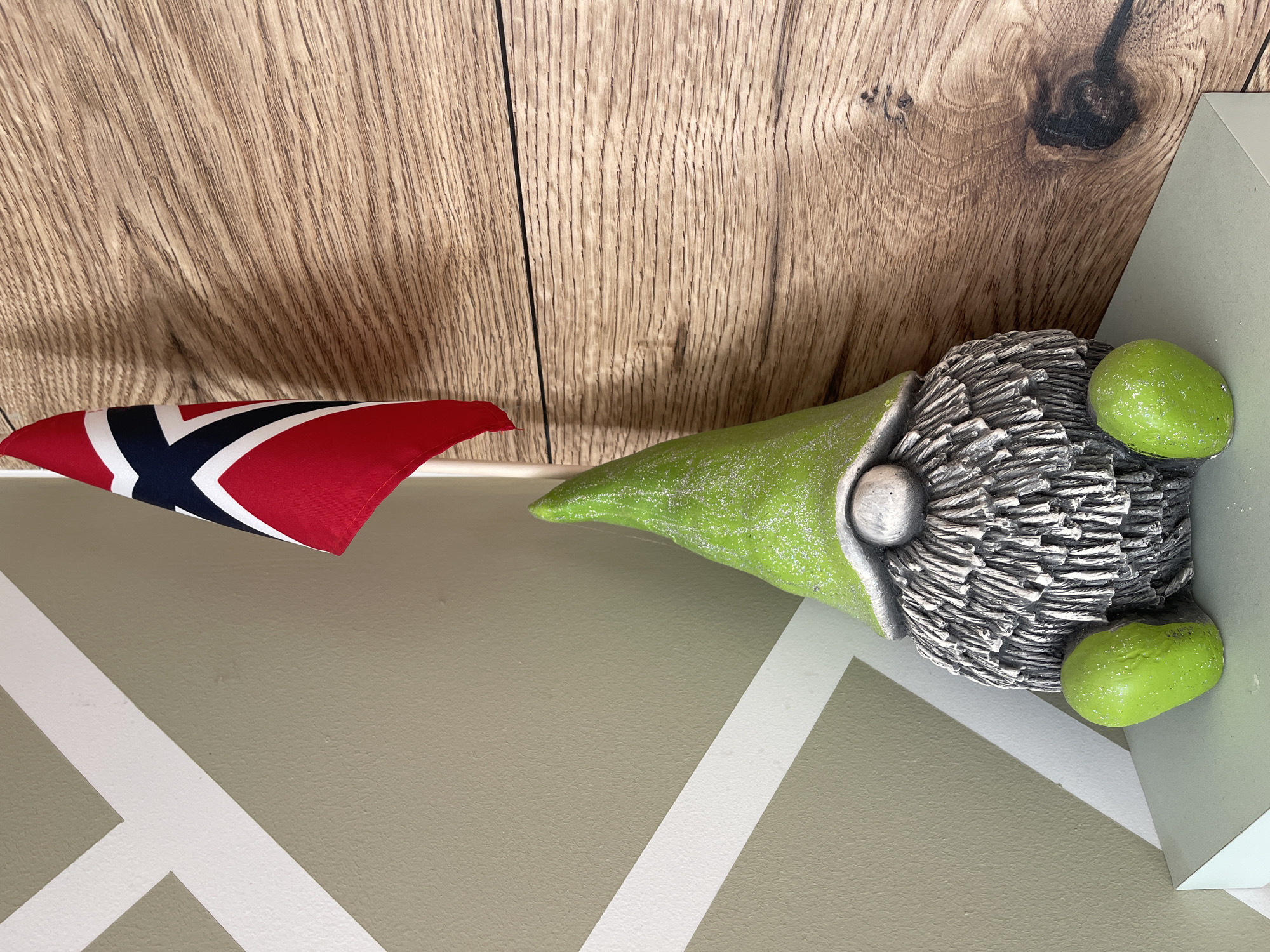 This image shows a garden gnome with a green hat and beard, wearing green shoes. A small Norwegian flag is attached to the gnome's hat. The gnome is sitting on a shelf in front of a wall with a geometric pattern and a wooden panel. The image was taken indoors, with no visible connection to Warsaw, Poland.