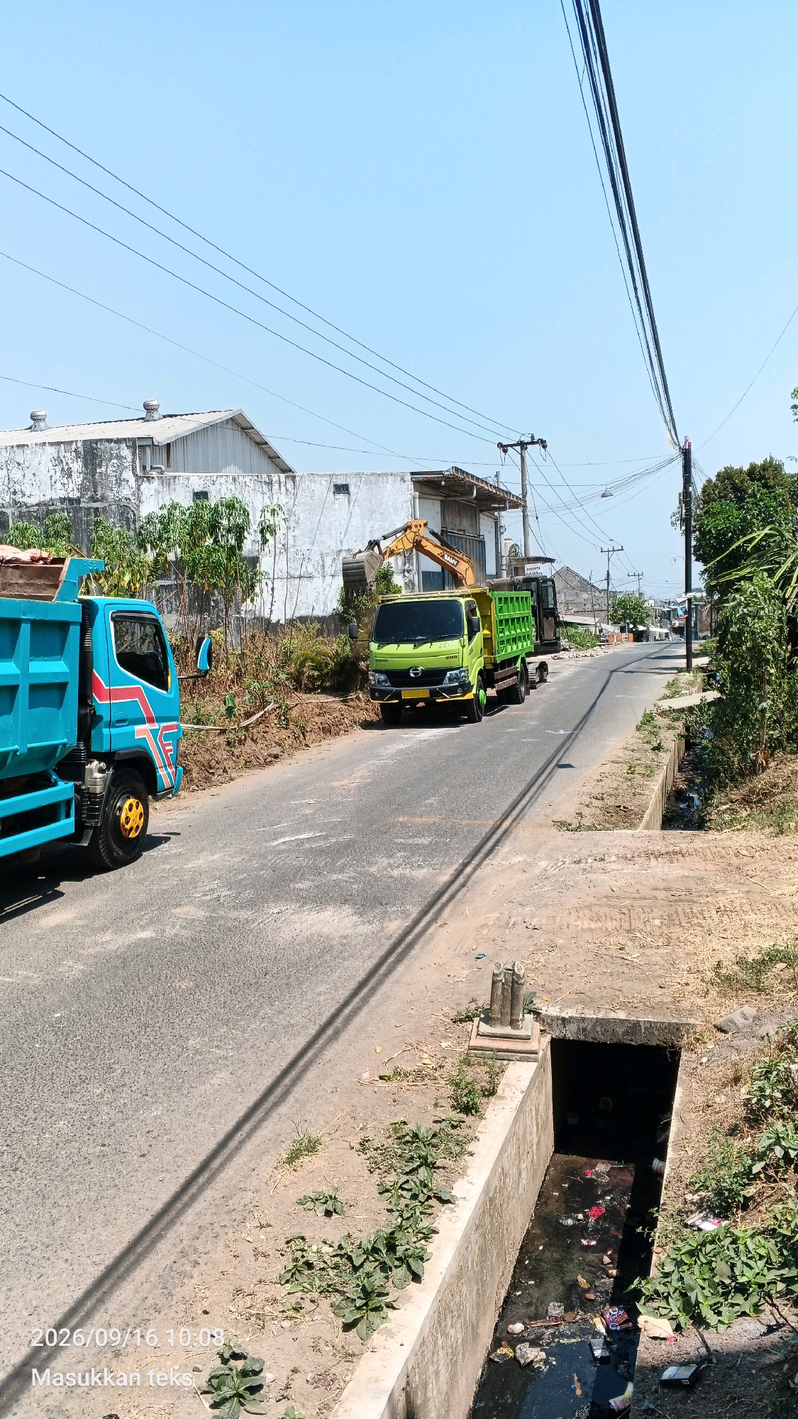In the city of Jeru, Indonesia, on September 16, 2026, at 10:08 AM, a street scene unfolds under a clear blue sky. The road surface shows signs of wear and tear, with patches of rough texture and scattered debris. To the left of the road, a bright turquoise dump truck with red and white accents is parked, its bed loaded with what appears to be dirt or gravel. Further down the road, a lime green dump truck is also present, facing away from the camera. This truck is equipped with a yellow excavator arm positioned above its cab.
Adjacent to the road on both sides are unpaved areas with sparse vegetation and dry grass. Buildings with weathered white exteriors are visible in the background, suggesting a residential or industrial area. Electrical wires crisscross the sky, supported by utility poles. A concrete drainage ditch runs along the right side of the road, filled with dark, murky water and visible trash, including plastic bags and wrappers. The scene appears to be a construction or road maintenance area, with the heavy machinery and materials indicating ongoing work. No people are clearly visible in the frame.