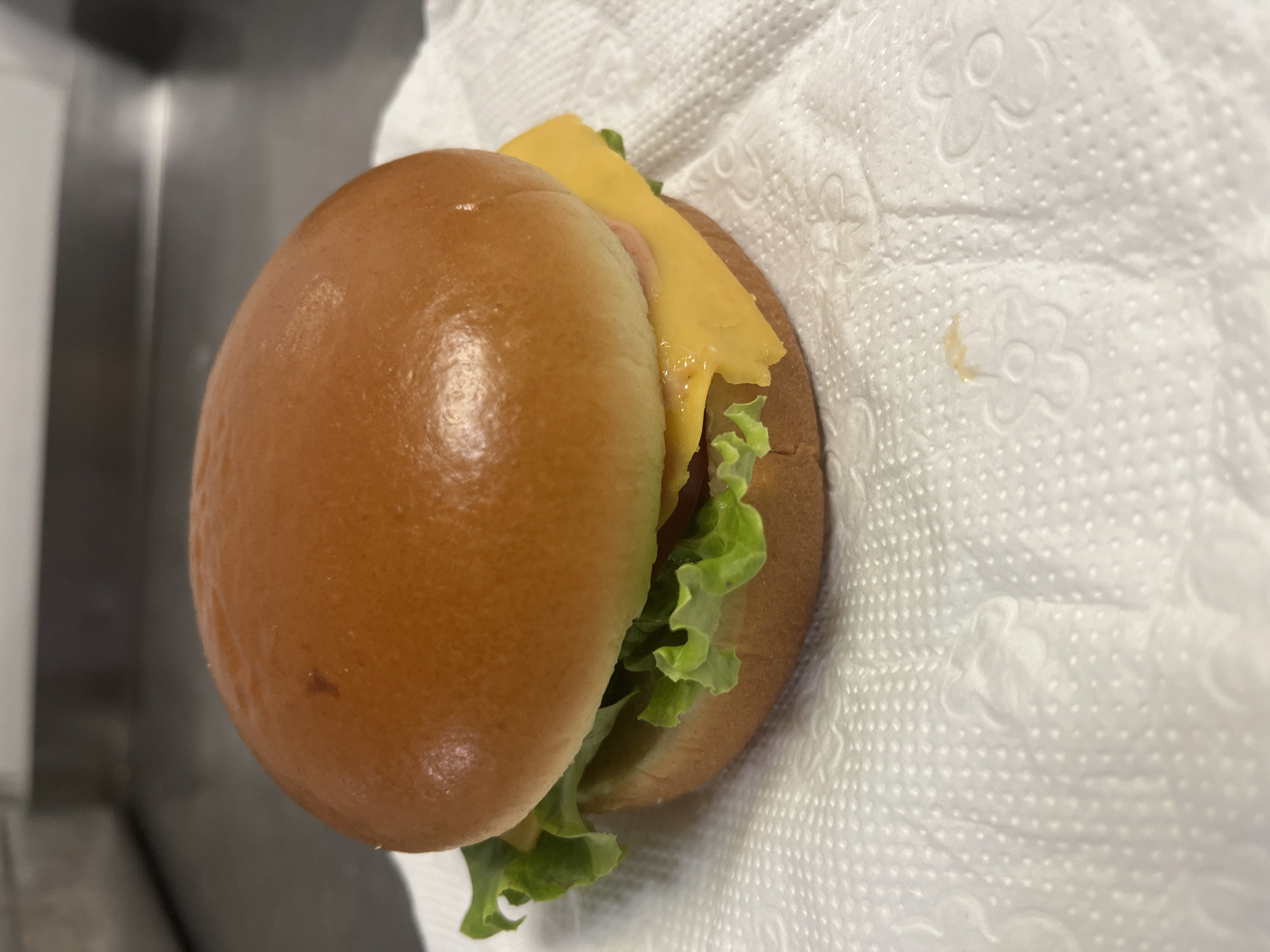 The image shows a close-up, overhead view of a small hamburger resting on a white, textured paper napkin. The hamburger is composed of a golden-brown bun, a slice of melted yellow cheese, a green lettuce leaf, and a hint of pink meat peeking out from under the cheese. The bun appears soft and slightly shiny. The napkin has a pattern of embossed floral shapes and a raised dot texture. The background is blurred, with a dark, vertical metallic surface visible on the left, suggesting an indoor environment, possibly a kitchen counter or a stainless steel surface. There are no people visible in the image, nor is there any text. The lighting is bright, and the focus is on the hamburger and the napkin. The image appears to capture a simple, prepared meal item.