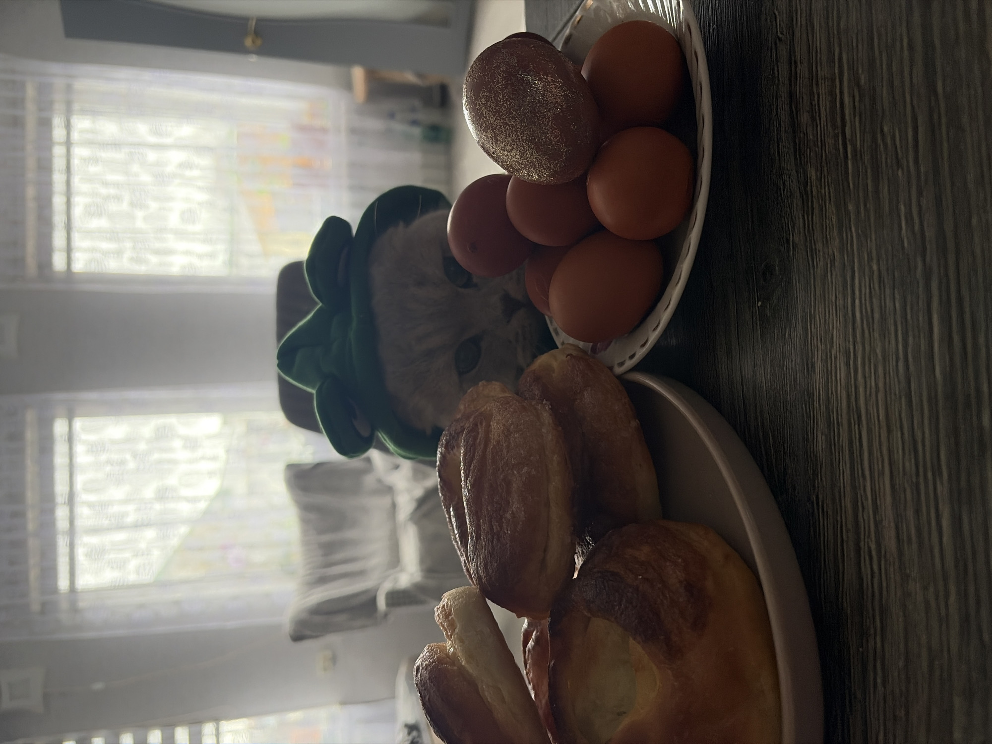 The image depicts a cat wearing a green hat, situated between two bowls filled with food. One bowl contains approximately seven brown eggs and one decorated egg. The other bowl contains baked goods. The scene is likely indoors, near a window. The location is Chaikovsky, Russia.