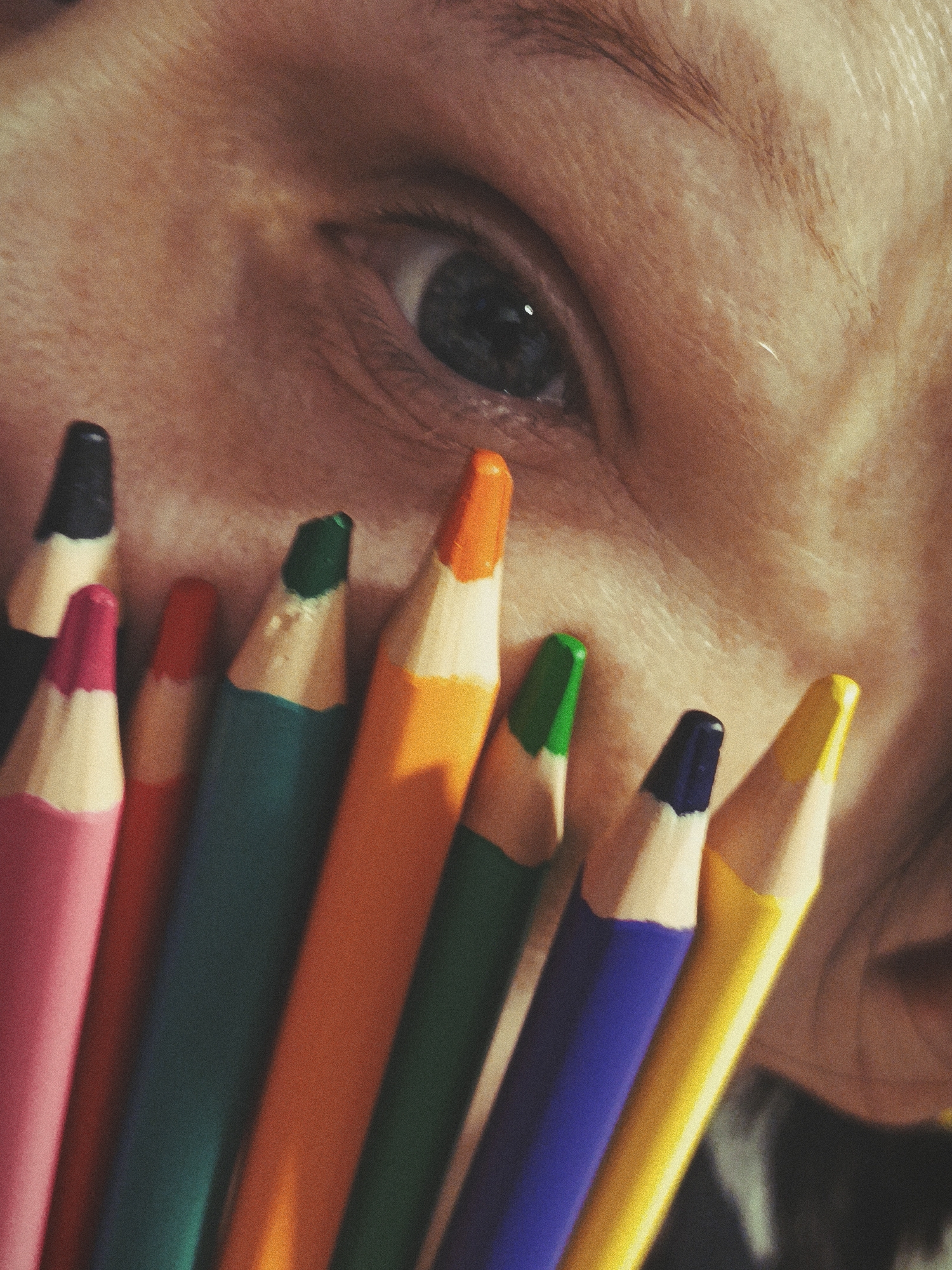 This close-up image features a human eye partially obscured by a collection of colored pencils. The eye, with a dark pupil and iris, is visible in the upper right quadrant, looking directly towards the viewer. The surrounding skin shows fine texture and some subtle creases, suggesting a tight crop of a face.
In the foreground, a vibrant array of colored pencils is arranged, appearing to rest against or be held close to the face. The pencils are positioned diagonally across the frame, showcasing their sharpened tips and wooden casings. Visible colors include pink, black, red-orange, dark green, bright orange, green, dark blue, and yellow. The pencils show slight wear, with some tips appearing used.
The image has a noticeable grainy texture, contributing to a somewhat soft, artistic aesthetic. The lighting highlights the pencils' colors and the contours of the skin. There is no visible text in the image, and no specific location, time of day, or weather cues are present within the frame. The composition creates an intimate and creative portrayal, blending a human element with artistic tools.