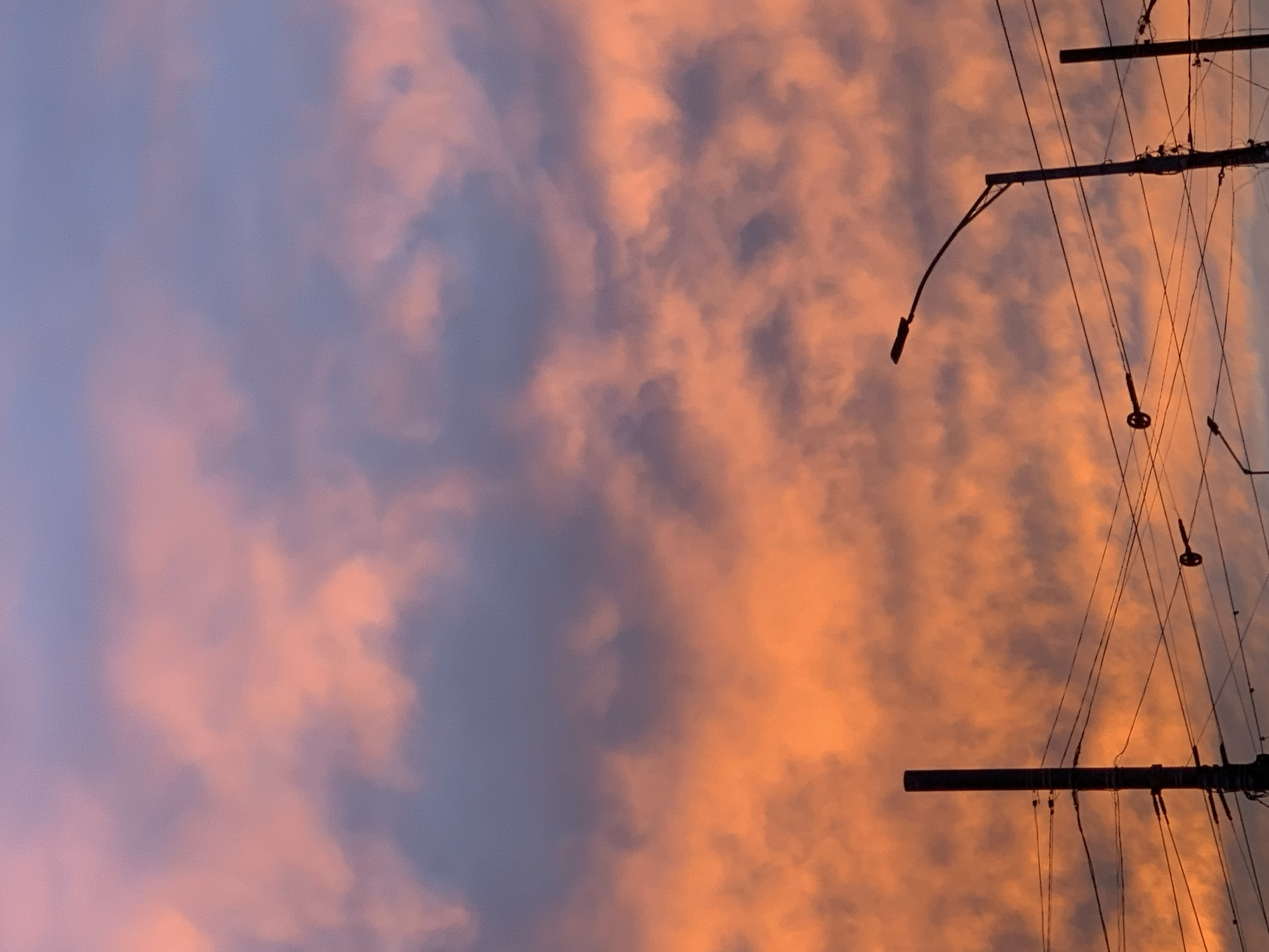 The image displays a vibrant sky at what appears to be sunrise or sunset, dominated by a striking blend of colors. The sky transitions from intense orange and peach hues on the right side to softer purples and blues on the left, with scattered clouds reflecting these warm and cool tones. The clouds vary in density, showing both diffused patches and more defined, streaky formations.
In the foreground, silhouetted against the colorful sky, are several utility poles and a dense network of overhead wires. These elements are concentrated primarily on the right side of the frame. One prominent street light fixture extends from a pole, curving downwards towards the left, with its lamp clearly visible in silhouette. Multiple horizontal and diagonal wires crisscross the frame, connecting the poles and appearing as dark lines against the bright sky. Other smaller, indistinct fixtures are also attached to the poles and wires.
The time of day is clearly either dawn or dusk, indicated by the dramatic and colorful lighting of the sky. The presence of utility infrastructure suggests an urban or suburban setting. No people are visible, and no specific activity or event is taking place beyond the observation of the sky. There is no visible text in the image.