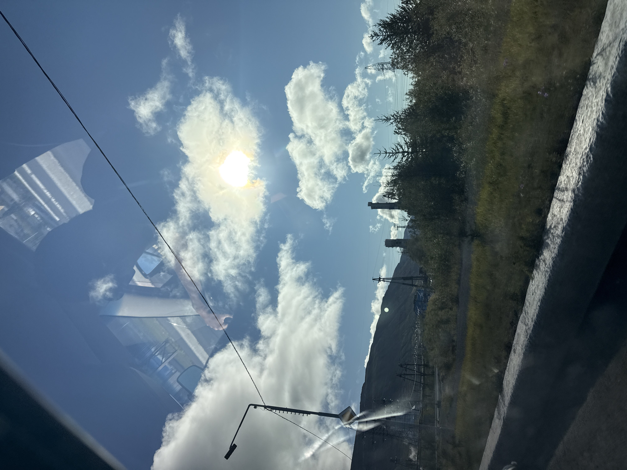 The image depicts an industrial landscape under a bright, cloudy sky, likely taken from a vehicle's window. The foreground features a concrete barrier and a grassy embankment. In the midground, a line of trees and vegetation is visible, interspersed with industrial structures such as power lines, tall poles with lights, and what appear to be factory chimneys or cooling towers. A mountain range forms the backdrop. The sky is a vivid blue with scattered white clouds, and the sun is shining brightly, creating lens flares and reflections on the glass. The reflections on the glass show a partial view of the interior of a vehicle, including a rearview mirror and the dashboard. The overall scene suggests an overcast but bright day in an area characterized by industrial activity and natural terrain, consistent with the environment of Norilsk, Russia, which is known for its mining and smelting operations and its proximity to mountains. There are no people clearly visible in the image.