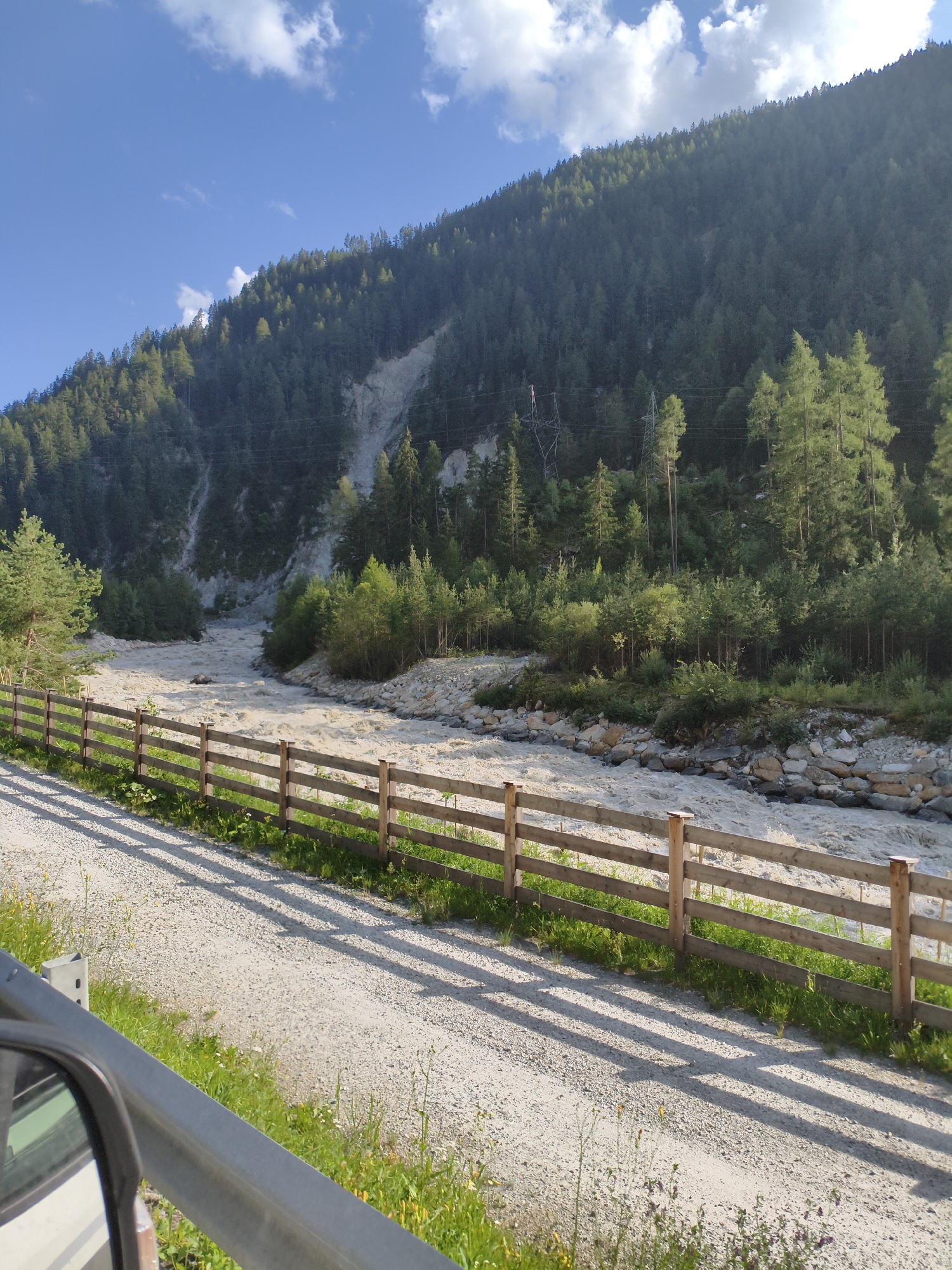 The image shows a wide, muddy river flowing through a valley in Köfels, Austria. The riverbanks are lined with stones and vegetation, and a dense forest covers the steep mountain slopes that rise behind the river. A wooden fence runs along the edge of a gravel path that parallels the river. In the foreground, a portion of a car's side mirror and railing is visible, suggesting the photo was taken from a moving vehicle. The sky is blue with scattered white clouds, and the sun appears to be casting shadows across the path. There are no people visible in the image. The scene depicts a natural landscape with signs of human presence, such as the fence and the path.
