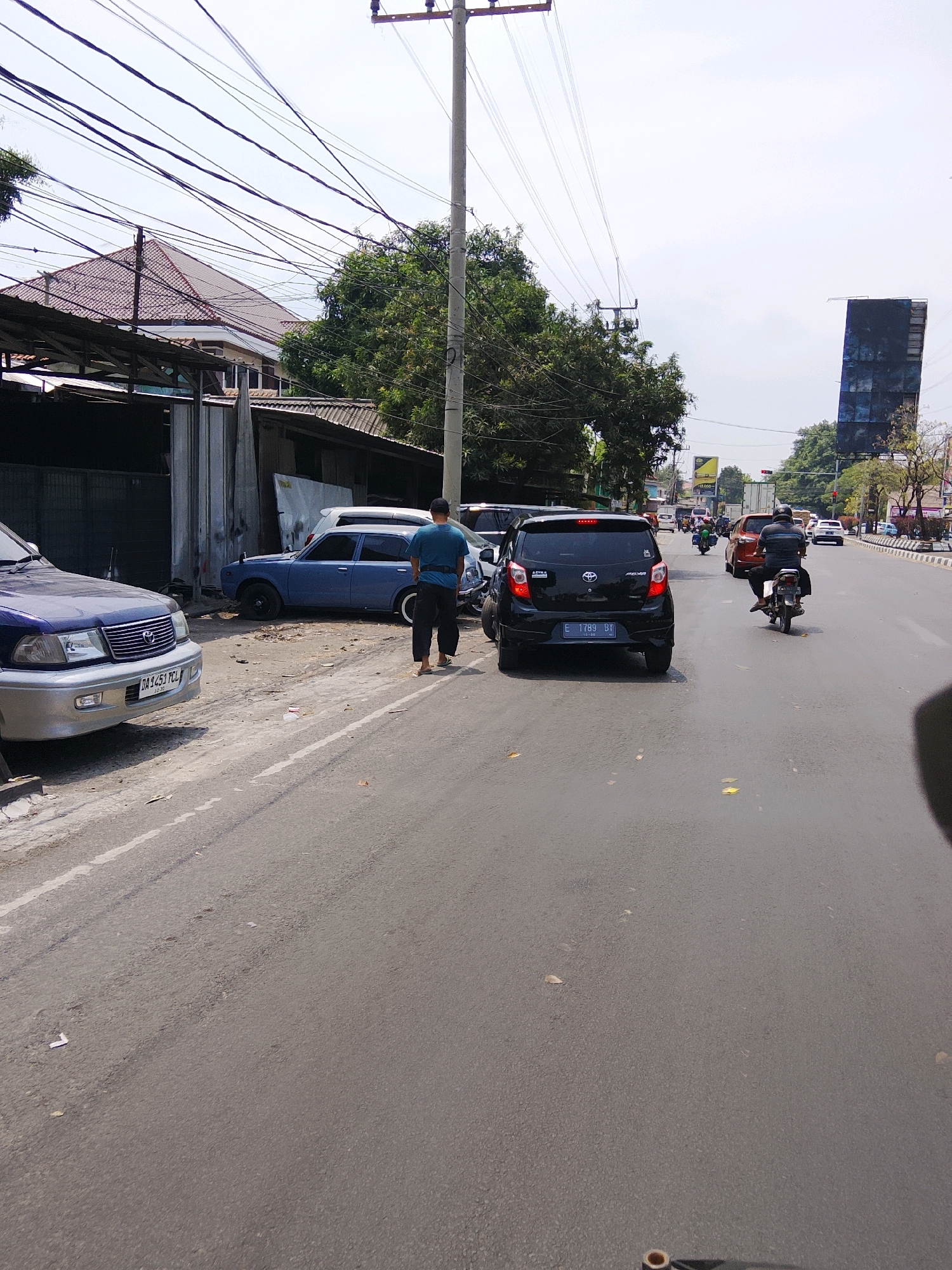 The image captures a street scene in Cirebon, Indonesia, under a bright sky. In the foreground, a blue sedan is parked on the left side of the road, its front bumper and headlights clearly visible. Further down the road, a person in a blue shirt and dark pants stands facing away from the camera, appearing to be near a blue car. A black hatchback is also parked on the road. Several other vehicles, including motorcycles and cars, are visible in the distance, suggesting moderate traffic. The environment includes buildings on the left with corrugated metal and tiled roofs, a utility pole with numerous wires, and trees. The road surface is asphalt with some scattered debris.