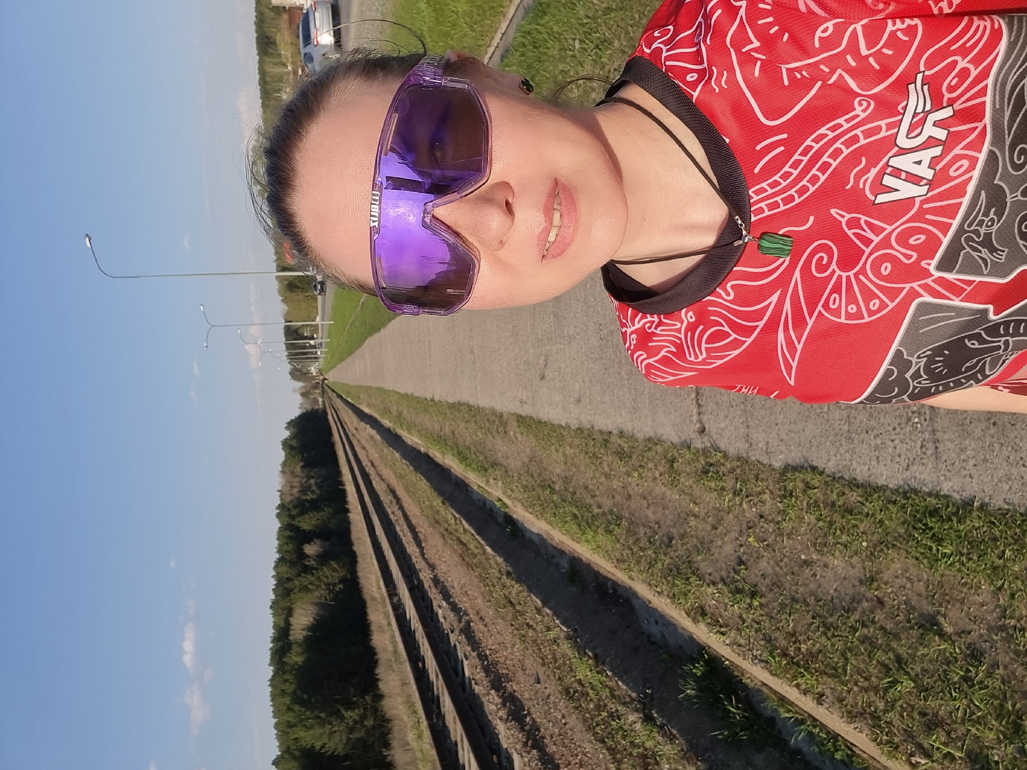 A waist-up shot shows a woman with pale skin wearing large purple sports sunglasses and a red athletic shirt with white and gray designs. She has dark hair pulled back and is wearing a black necklace with a small green pendant. The background shows railroad tracks extending into the distance, with green foliage on either side. A paved path runs alongside the tracks, and streetlights are visible along the road in the background. The sky is blue and clear. The scene is in Chaikovsky, Russia.