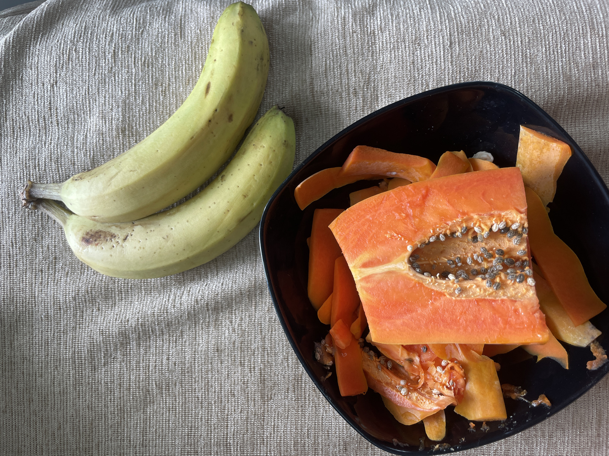 Two yellow bananas are situated on a textured, light brown, woven surface. To the left of the bananas, a black bowl contains cut pieces of papaya, including a large slice revealing its seeds. The papaya pieces are a vibrant orange color. The overall scene is lit from above, creating soft shadows. There is no indication of the city of Bhagsu, India.