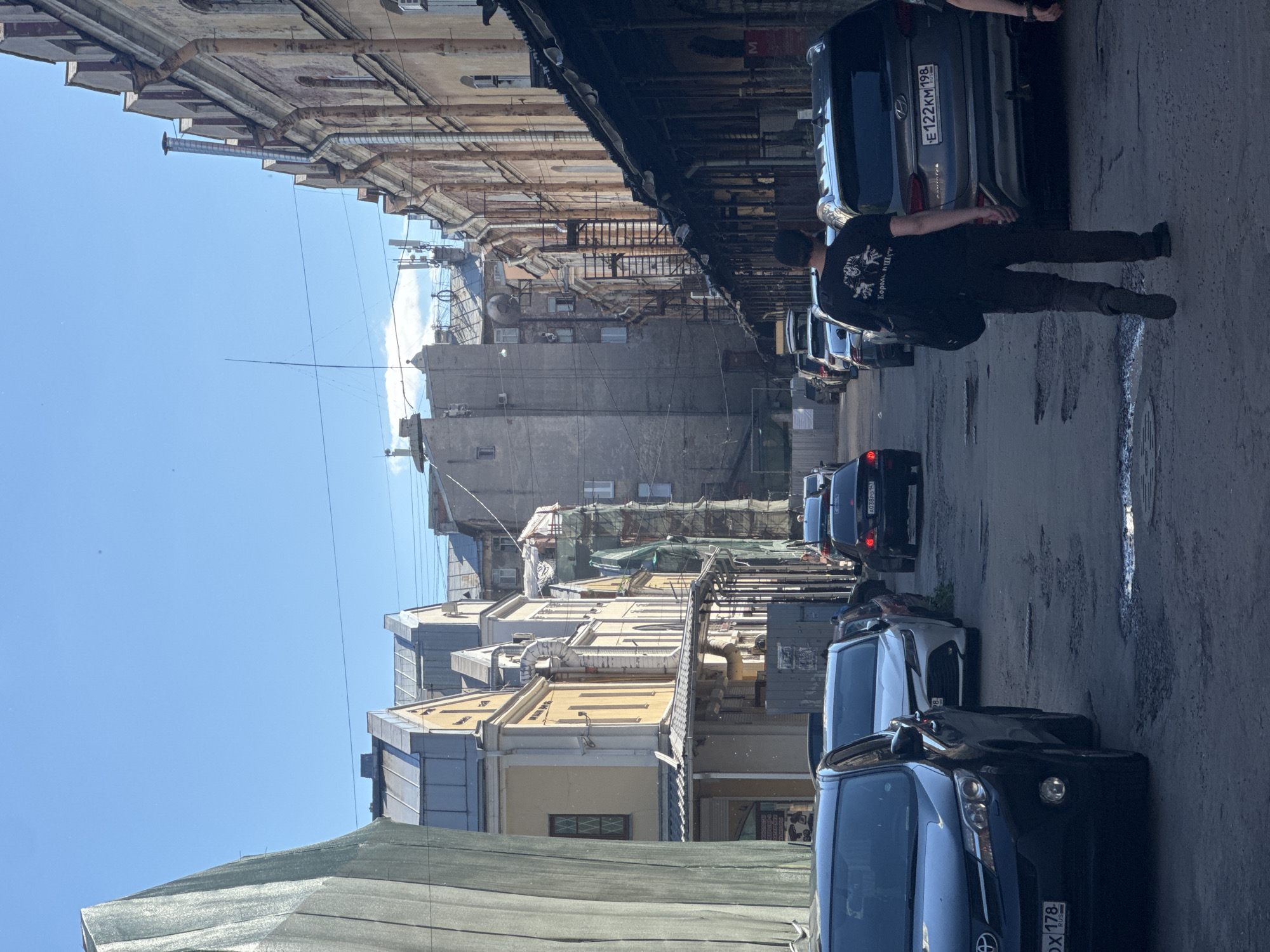 The image depicts a narrow street in Saint Petersburg, Russia, lined with historic buildings. The street is paved with asphalt and shows signs of wear and tear with several potholes. Cars are parked on both sides of the street, and some are in motion. A man wearing a black t-shirt with graphics and dark pants is walking away from the camera, carrying a backpack over his left shoulder. The buildings on the right have a covered walkway or arcade along the ground floor. On the left side of the street, there's a building covered with a large section of green protective netting, suggesting ongoing construction or renovation. The sky is clear and blue, indicating a sunny day. The architecture suggests a historical district of the city.