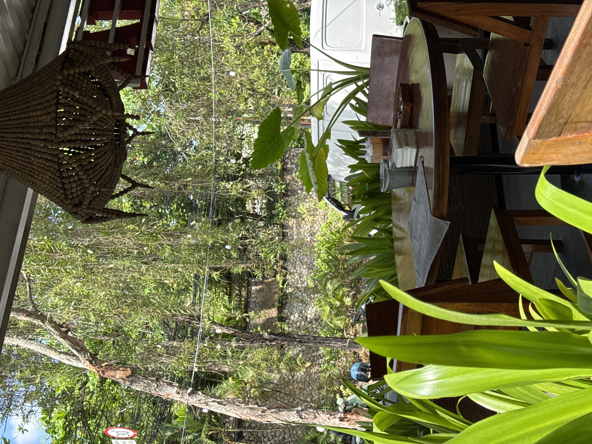 This image captures a scene at a restaurant in Kuta Selatan, Indonesia. Wooden tables and chairs are set on a patio, with lush greenery providing a backdrop. A decorative light fixture hangs overhead, and the restaurant appears to be open-air. The surroundings suggest a tropical environment, with a stone pathway and dense foliage visible in the distance. There are plants decorating the tables.